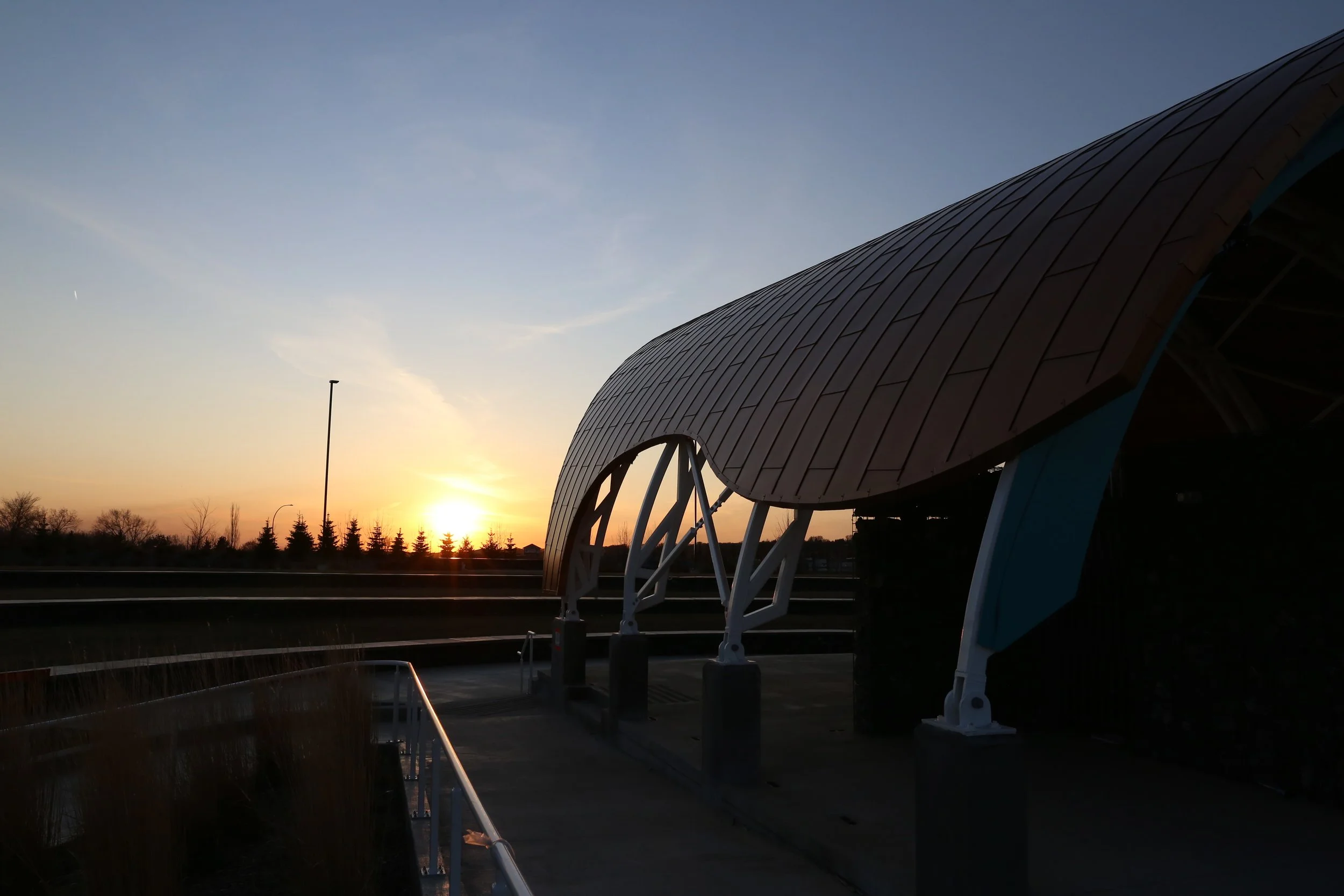 Sunset over a modern building with a curved wooden roof and white structural supports, near a paved walkway and a streetlight in the distance.