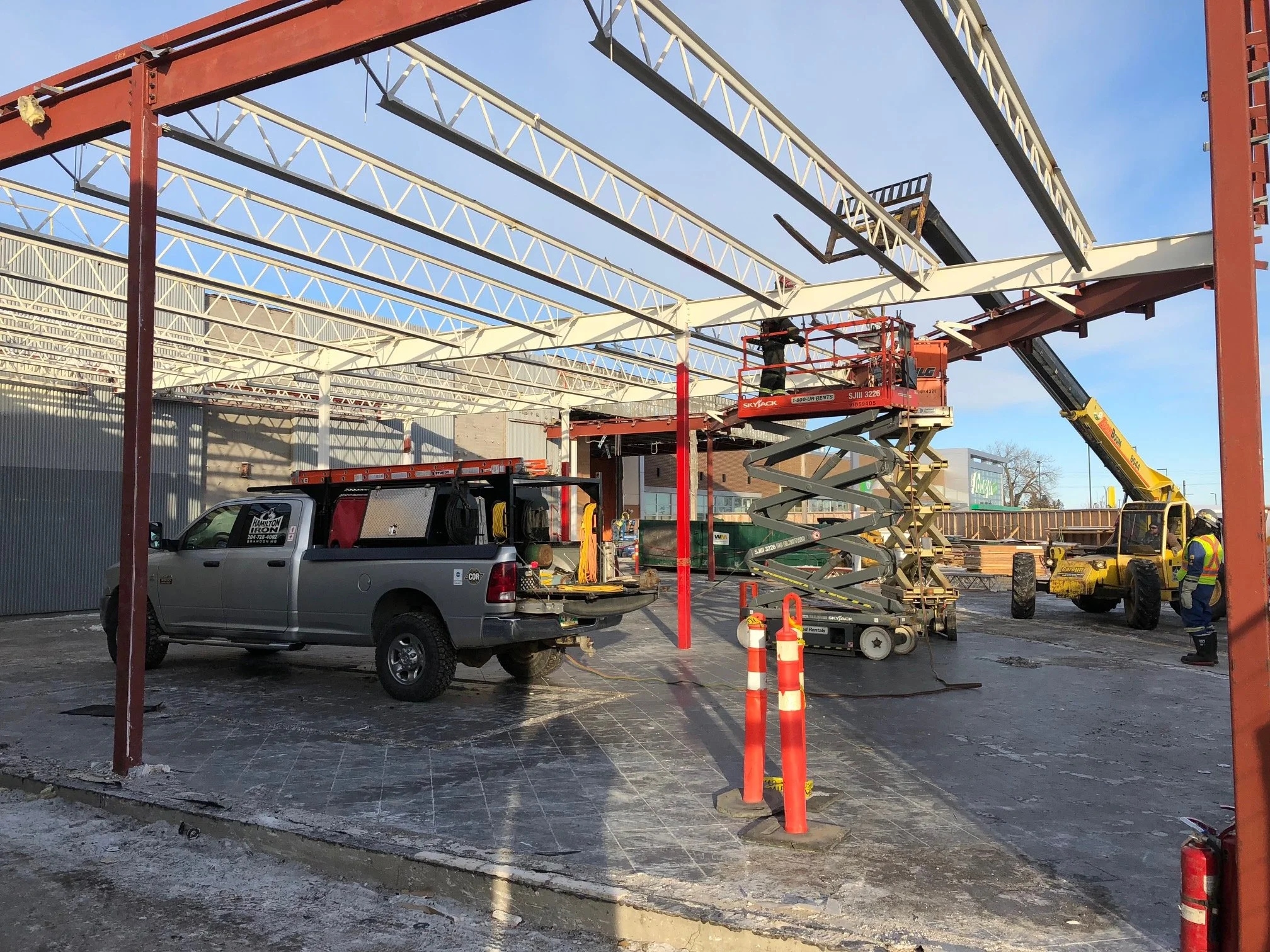 Construction workers and machinery assembling a metal framework of a building, with a work truck and a crane present on site.
