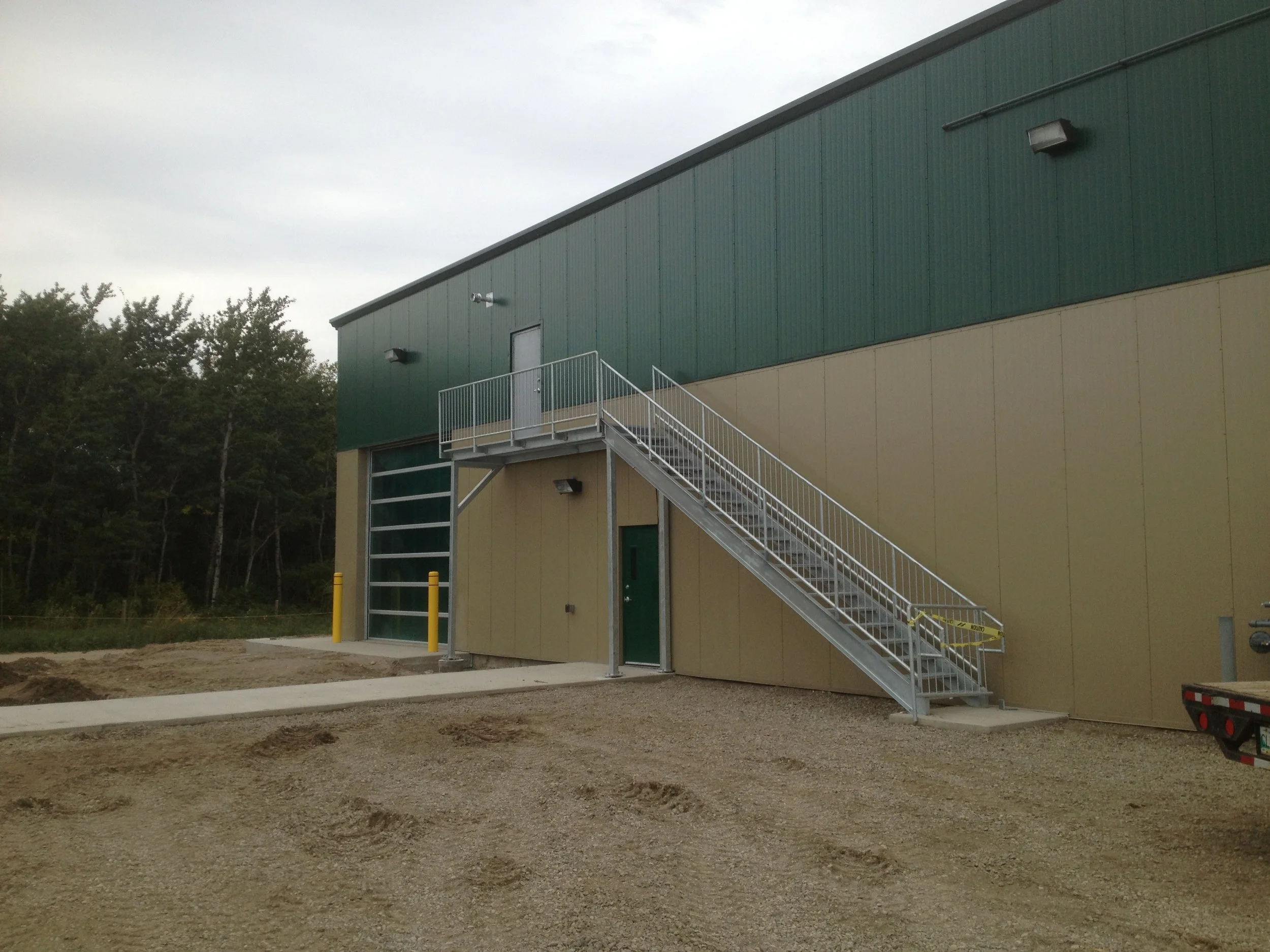 Exterior of a large industrial building with a metal staircase leading to an upper door, surrounded by a dirt lot with tire tracks and trees in the background.