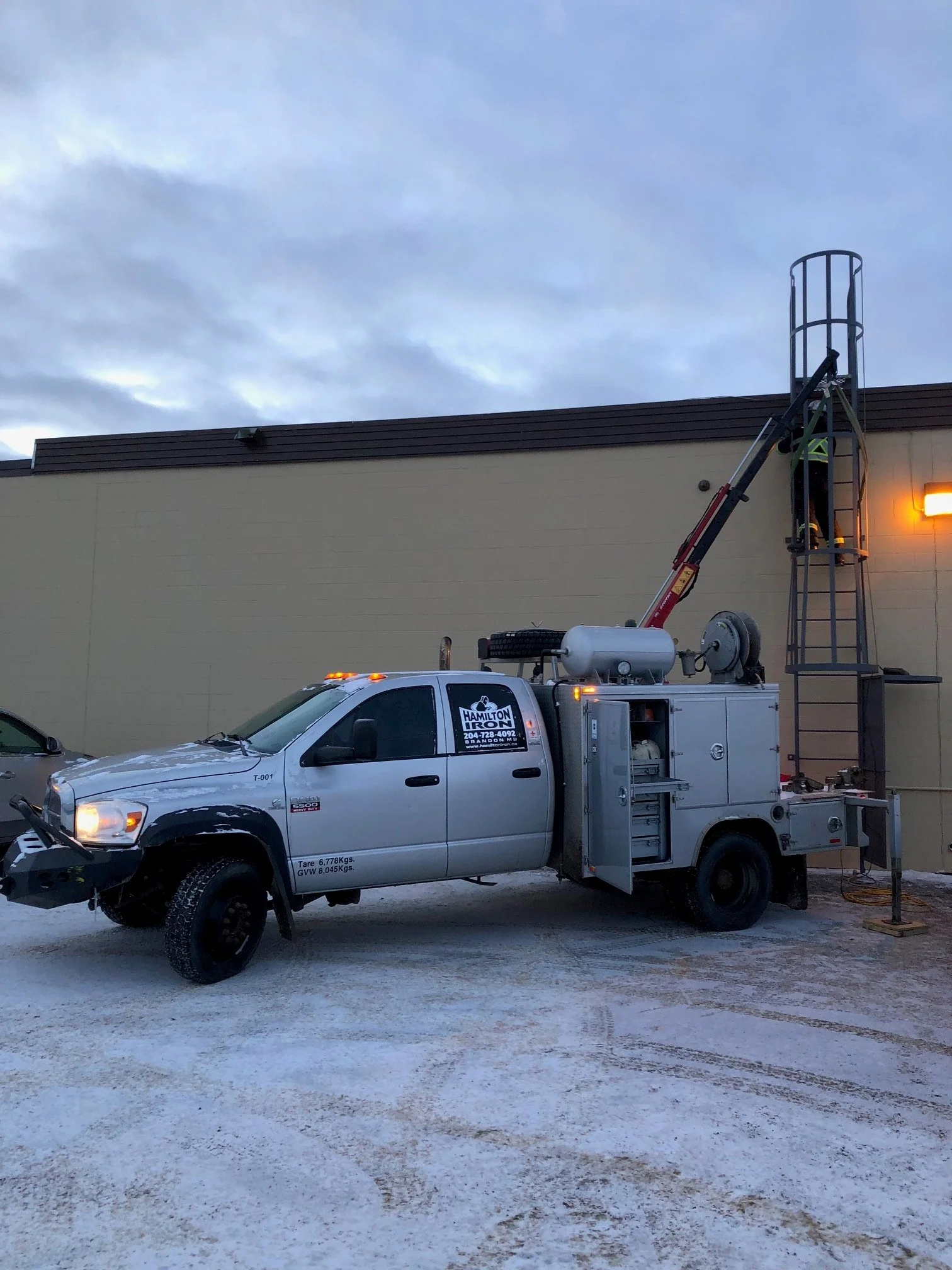 Utility truck with a worker on a tall ladder installing or repairing a light on a building exterior at dusk, with a cloudy sky overhead.