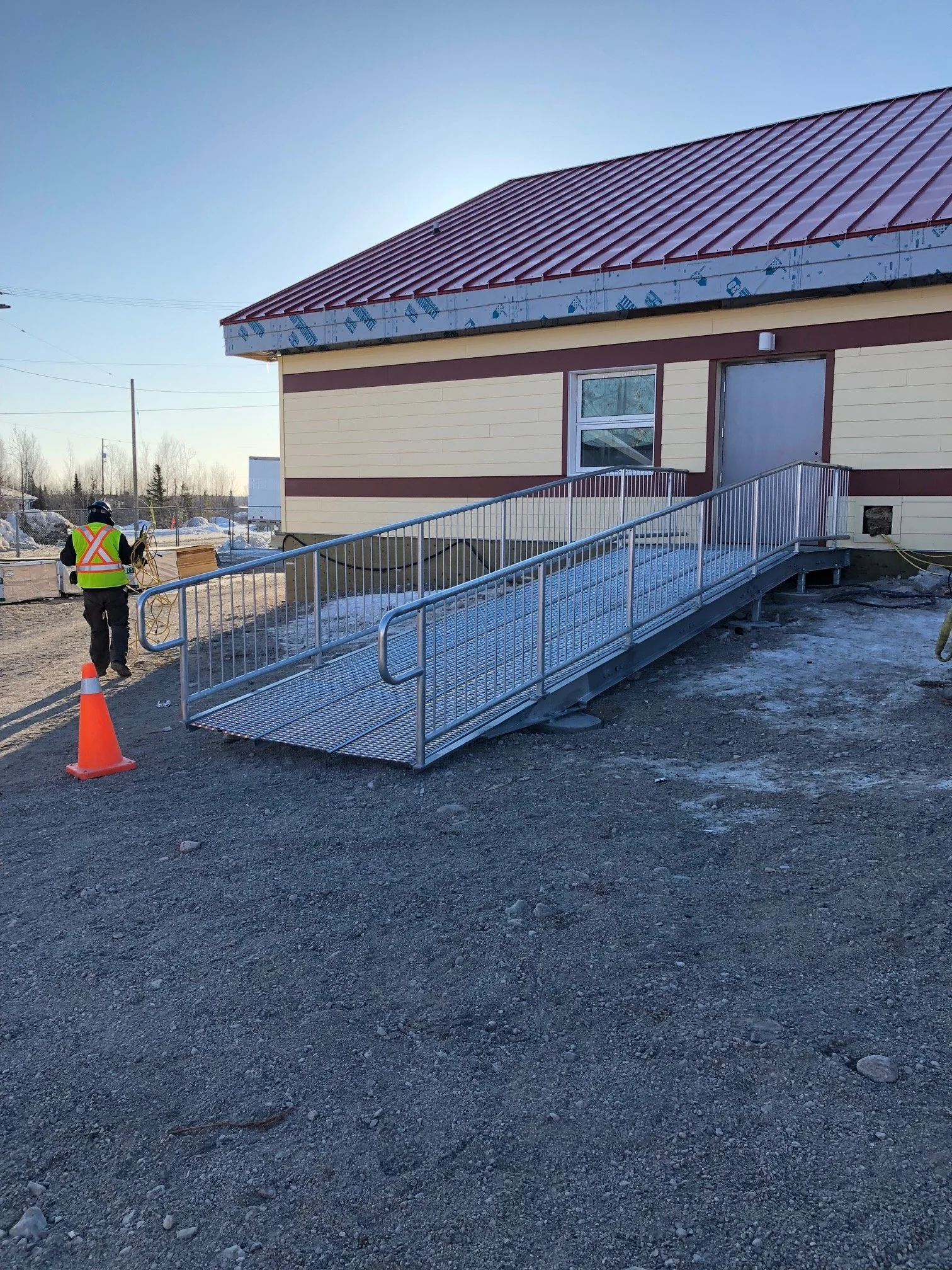 Construction site with a new metal wheelchair ramp leading to a building entrance, a construction worker in safety gear, and a safety cone on a dirt ground.