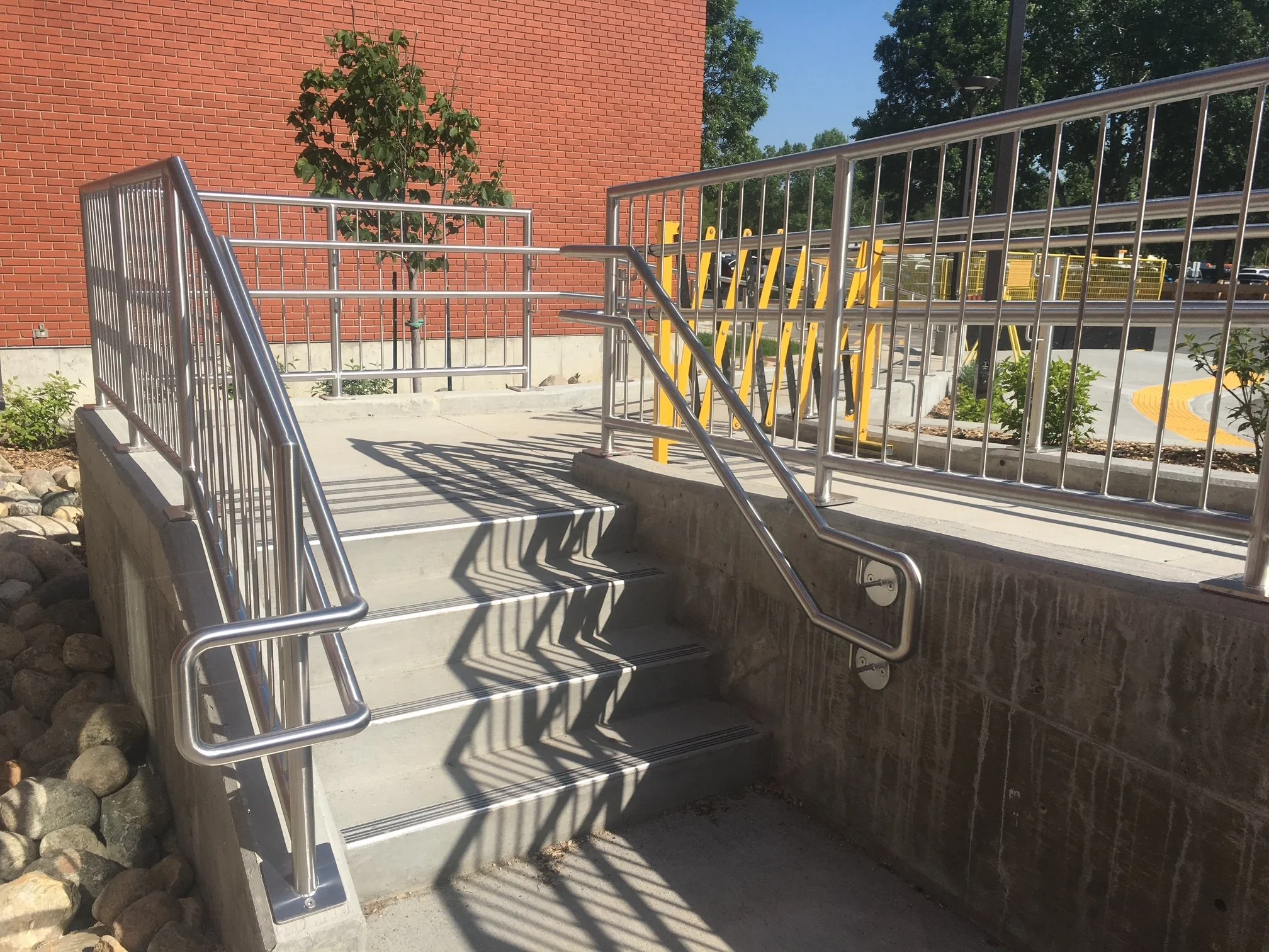 Metal wheelchair accessible ramp with handrails leading up to a sidewalk next to a brick wall, with a small tree and plants in the background.
