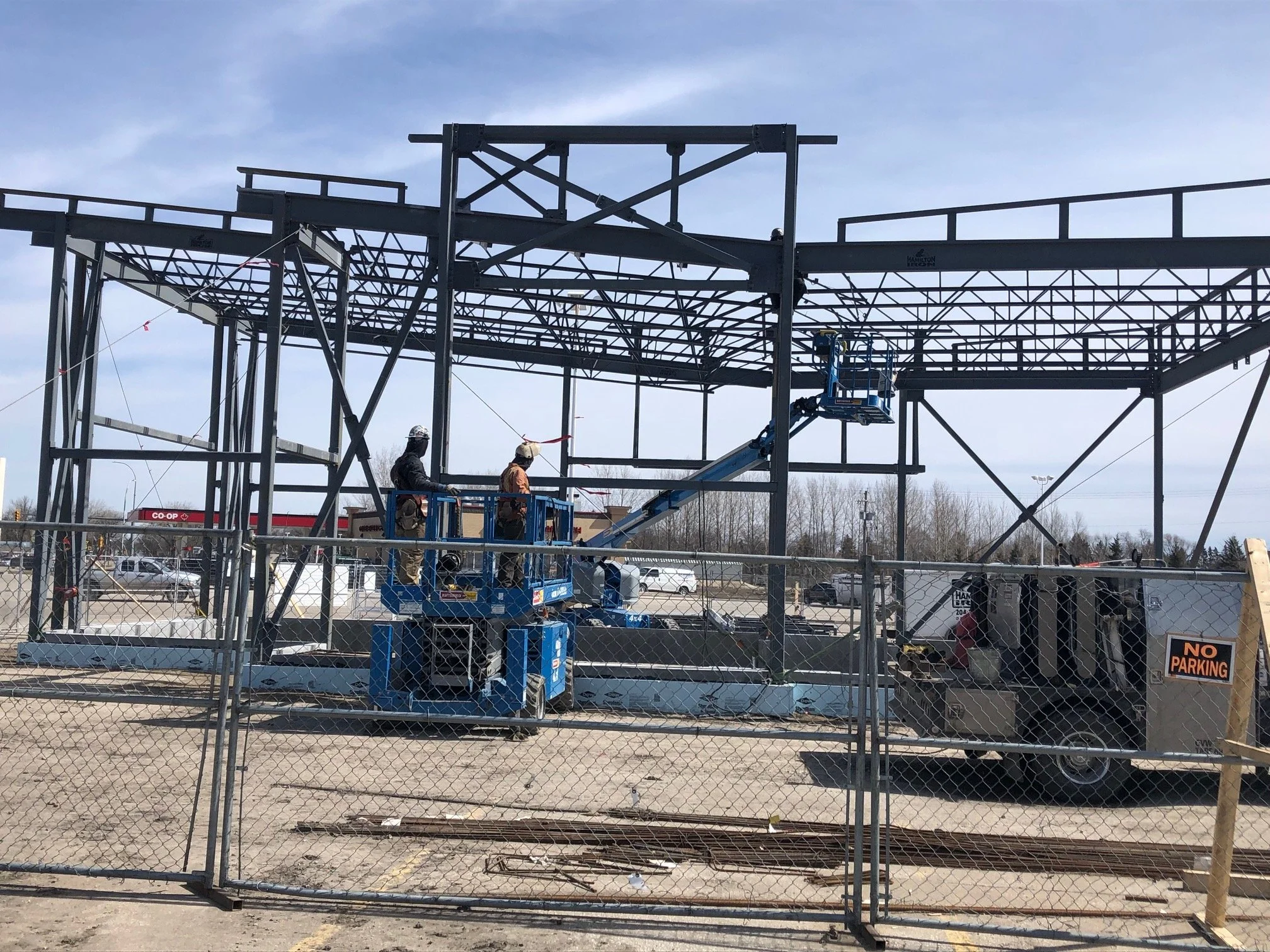 Steel framework construction site with two workers on a blue lift inside a fenced area, and a trailer with equipment nearby, under a cloudy sky.