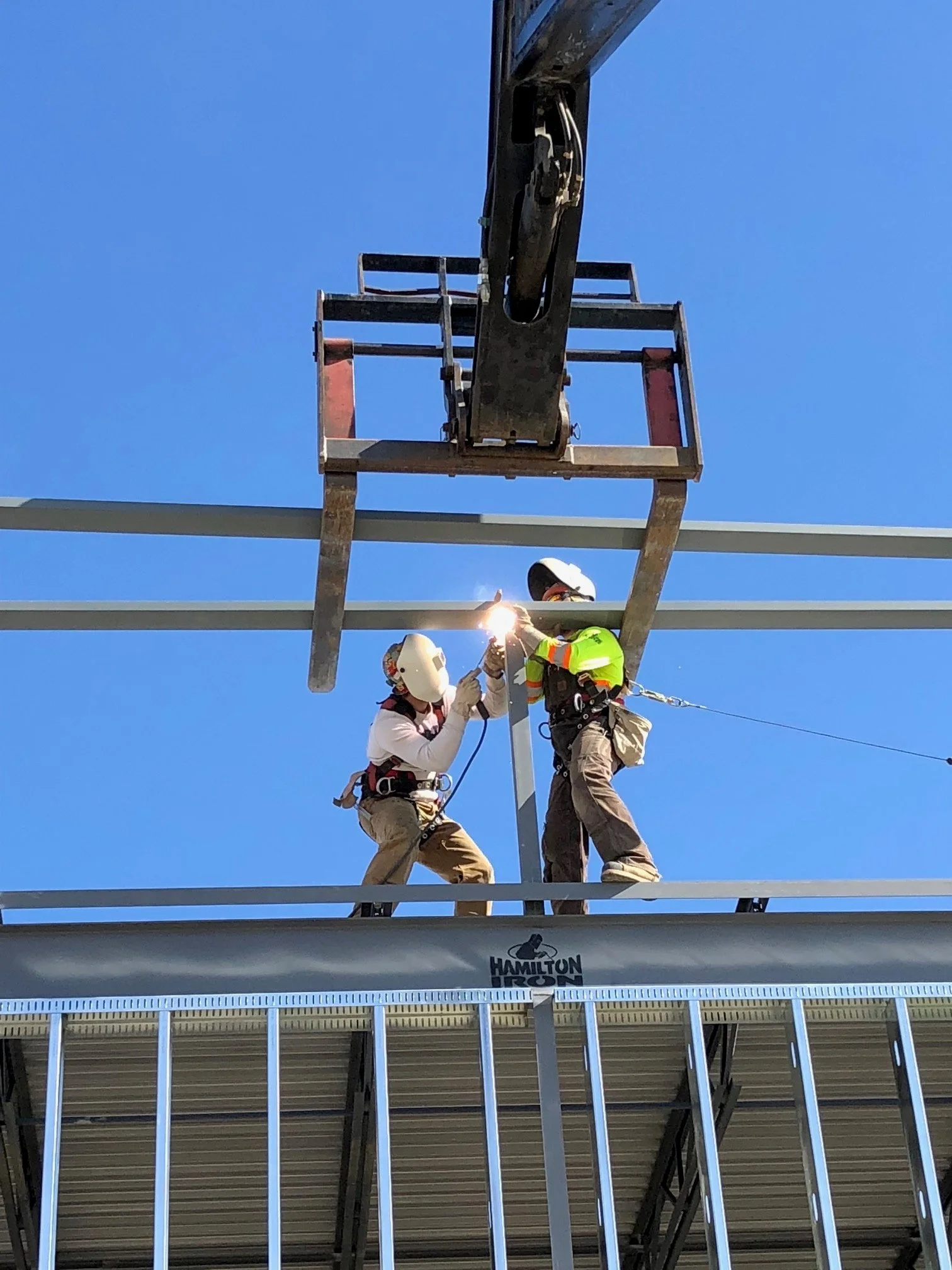 Two construction workers wearing safety helmets and harnesses working on a steel structure, with one using welding equipment and sparks flying, under a clear blue sky.