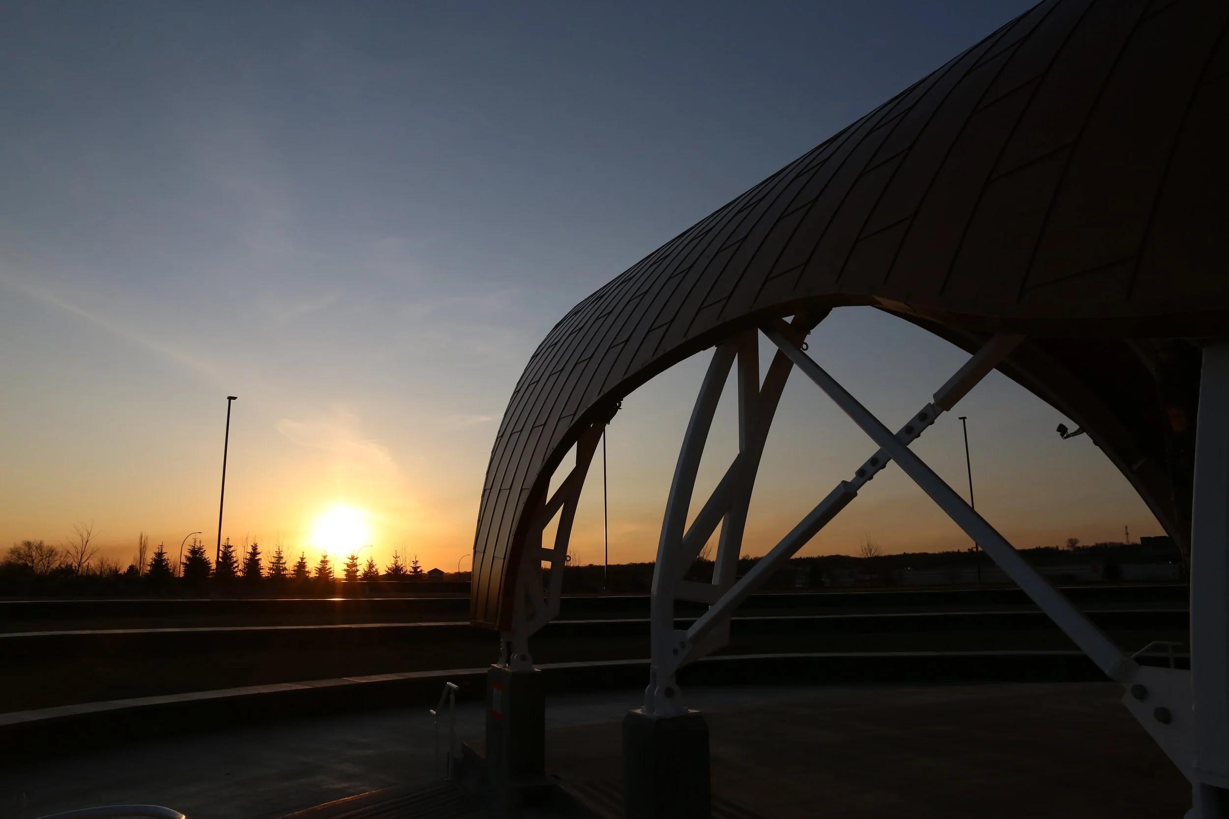 Sunset behind a modern, curved architectural structure with steel supports, on a road with trees in the distance.