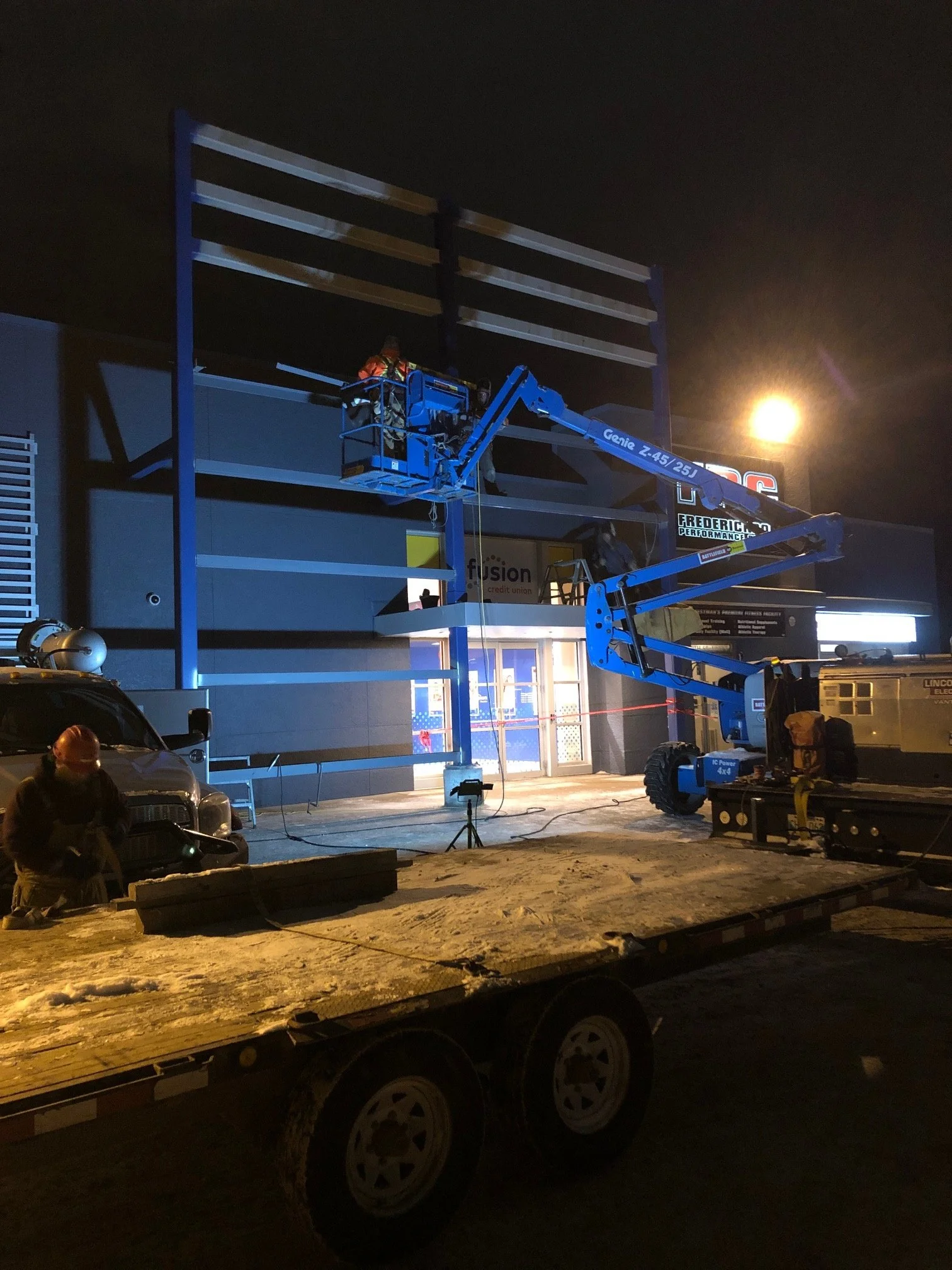 Workers installing signage on the exterior of a building at night using blue boom lifts.
