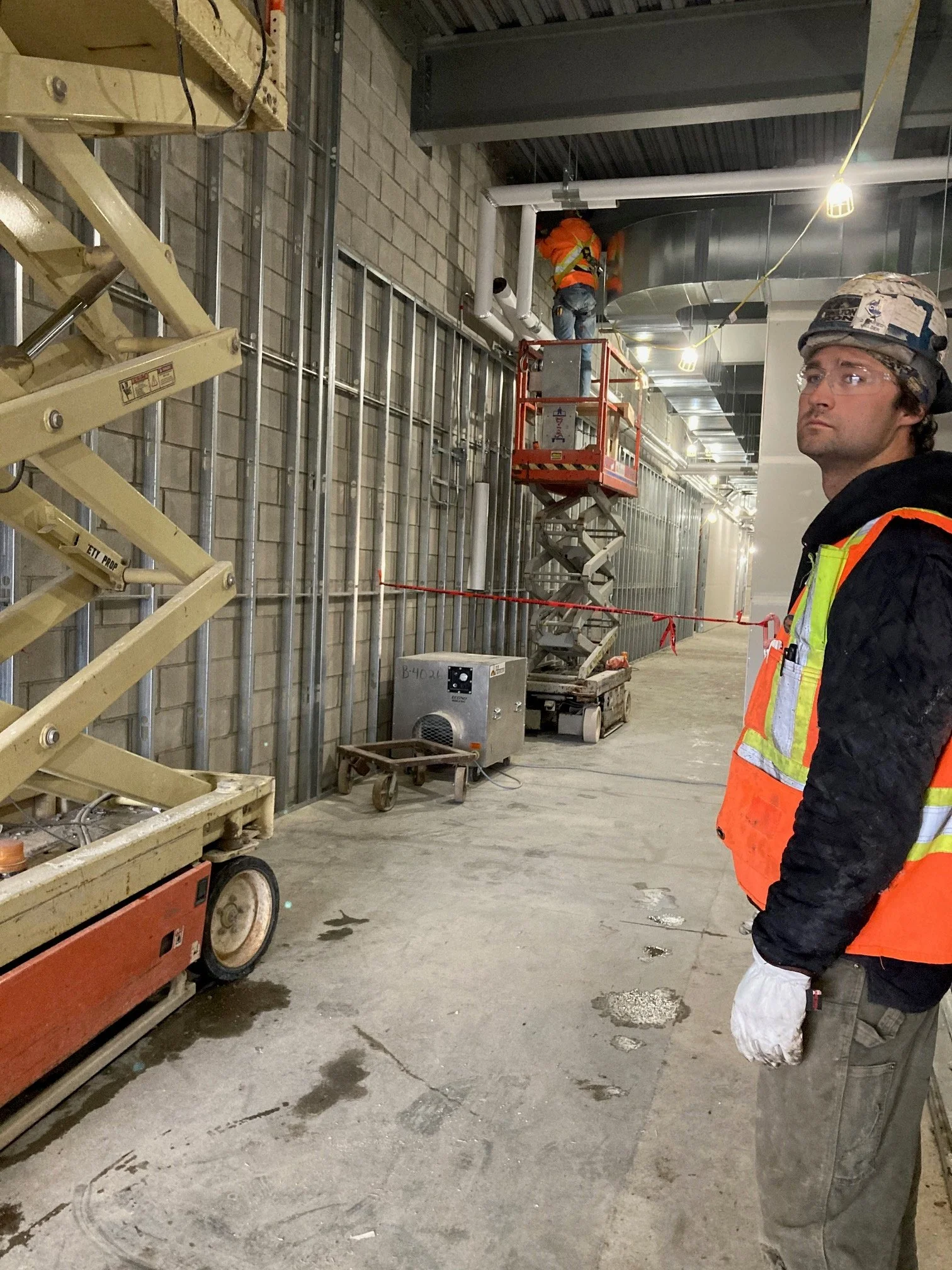 Construction workers in safety gear working inside a building under construction, with scaffolding, equipment, and exposed walls.