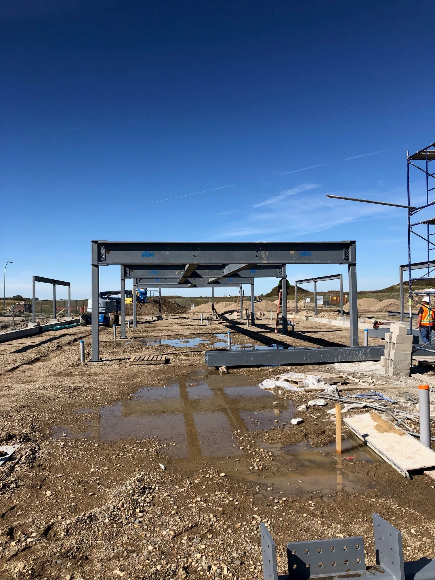 Construction site with steel framework and workers, muddy ground with puddles, clear blue sky in the background.