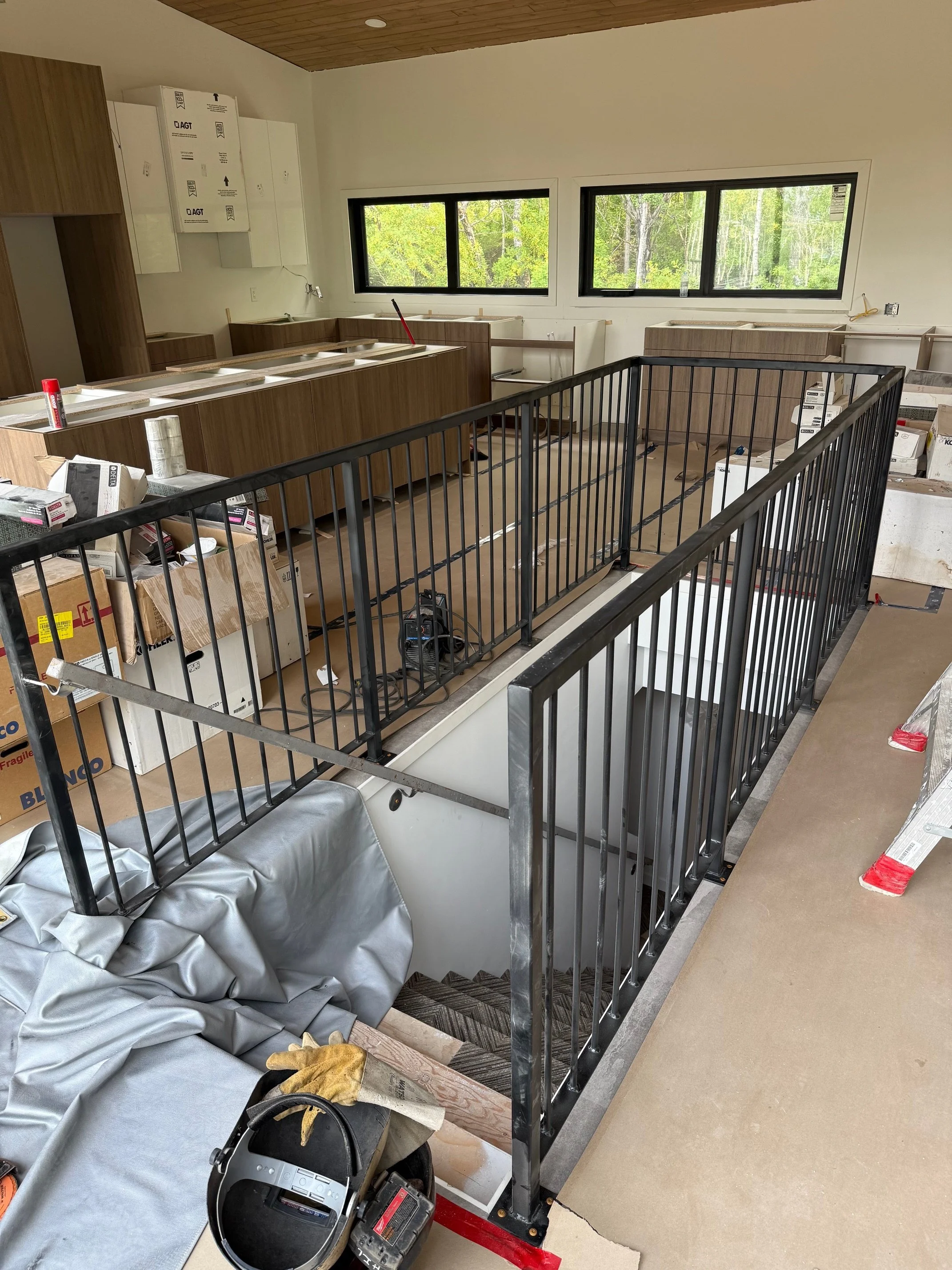 Interior of a house under construction, showing a staircase with black metal railing, unfinished cabinetry, construction tools, and materials scattered around.