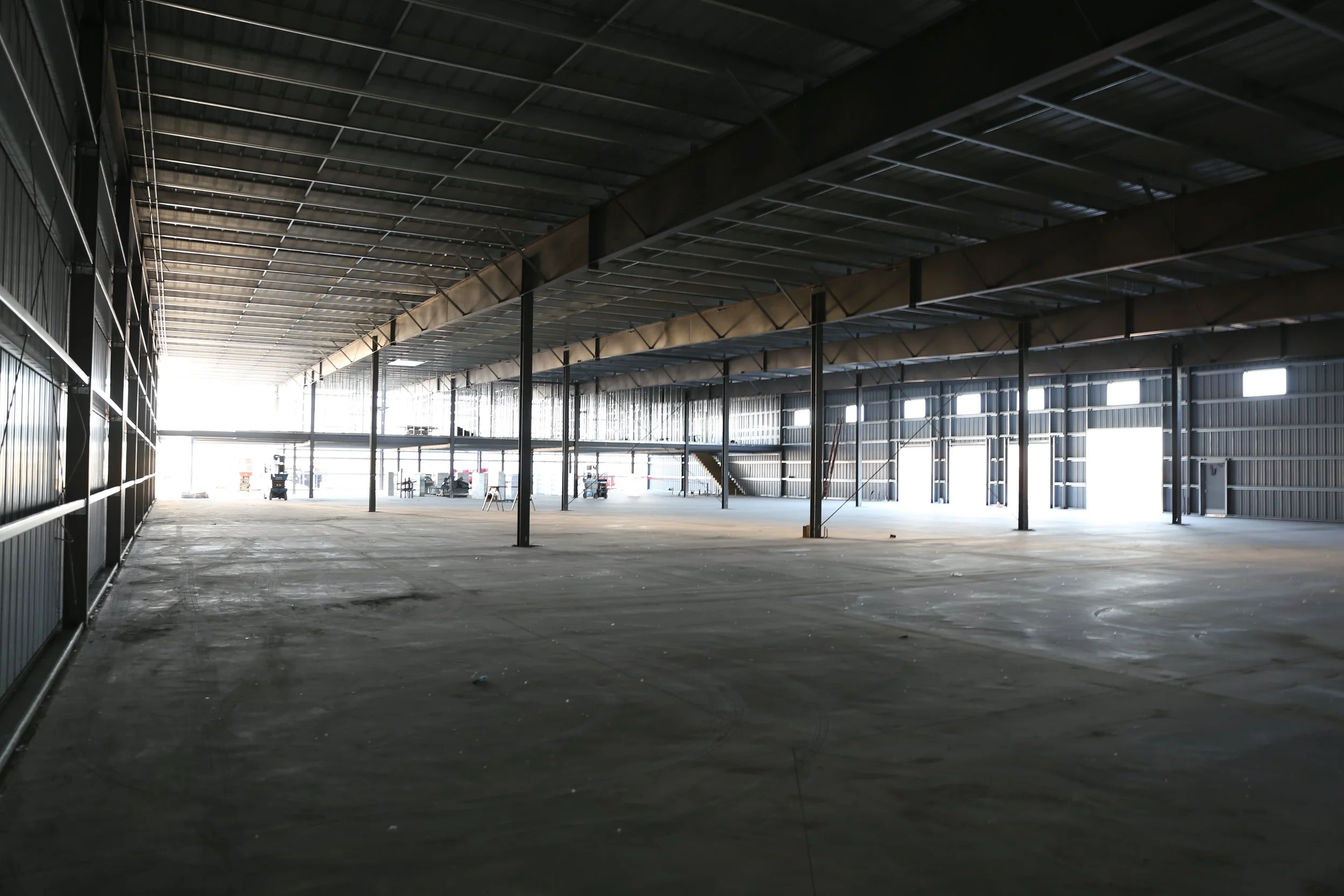 Empty warehouse with metal walls and ceiling, supported by black steel beams, with a polished concrete floor and bright natural light coming from open doors at the far end.