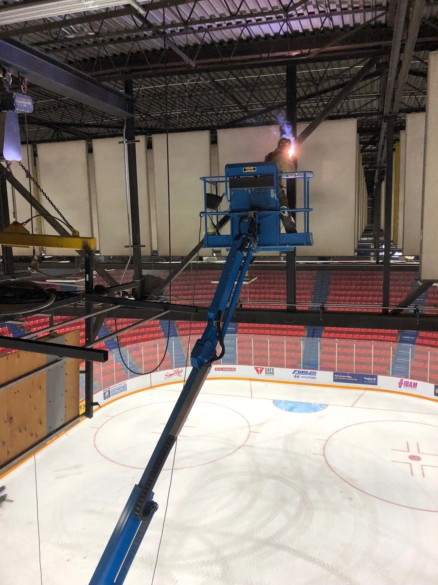 A worker in a blue lift is welding on a structure above an ice hockey rink, emitting bright sparks and smoke.