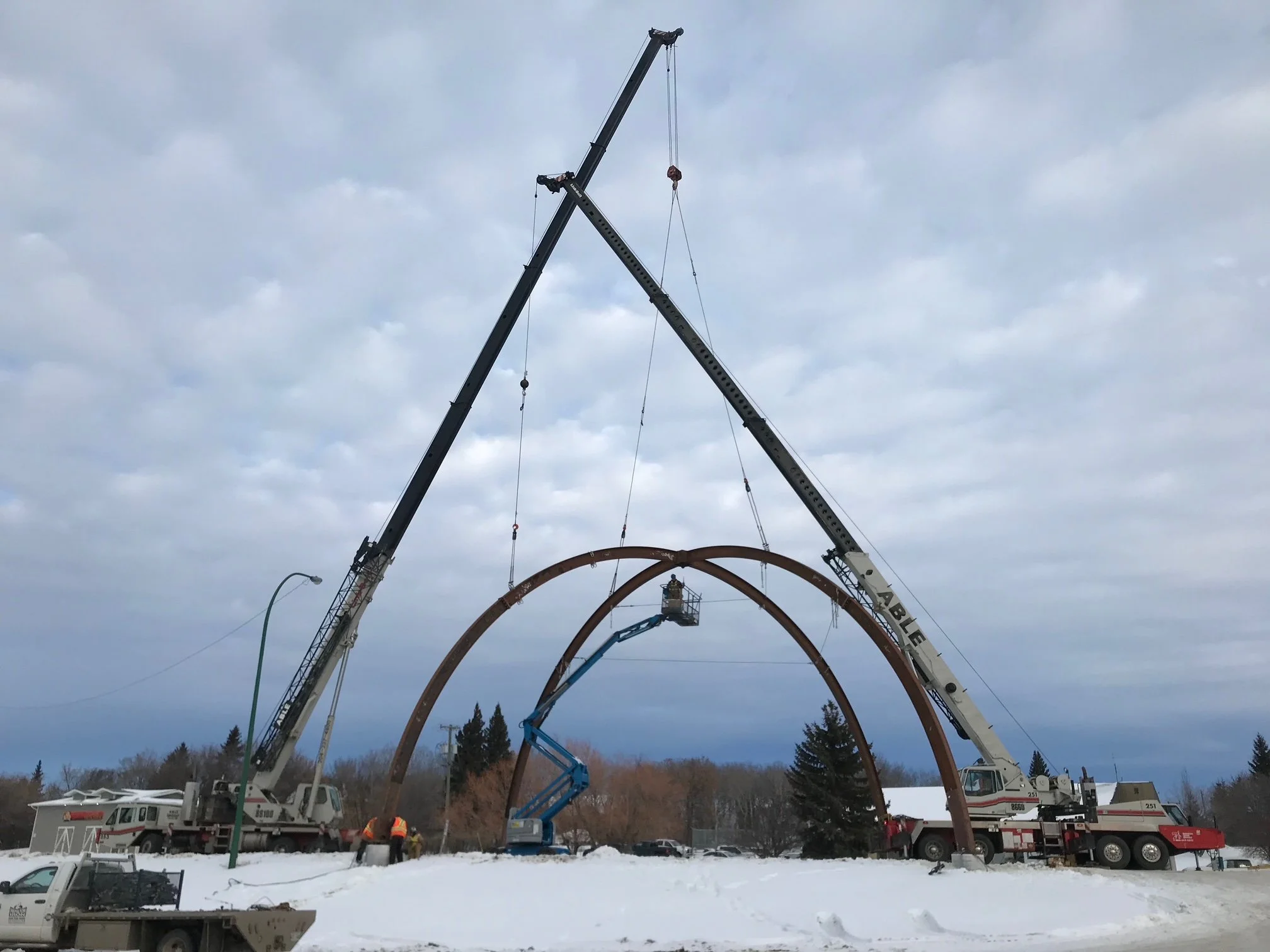 Two large cranes lifting a large, rust-colored metal arch structure in a snow-covered outdoor setting, with workers and trees in the background.