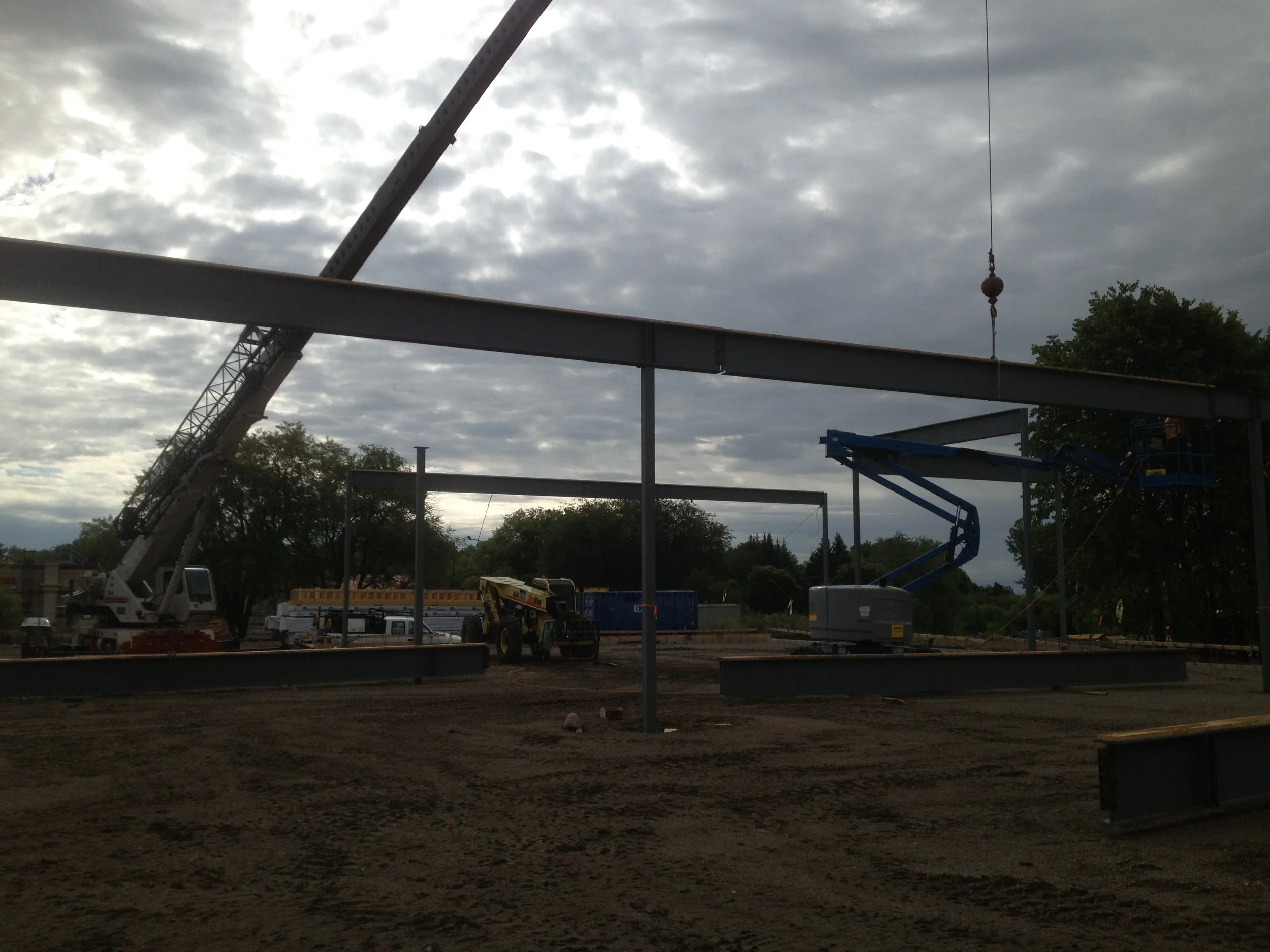 Construction site with steel beams being assembled, a crane, a boom lift, and construction equipment under a cloudy sky.
