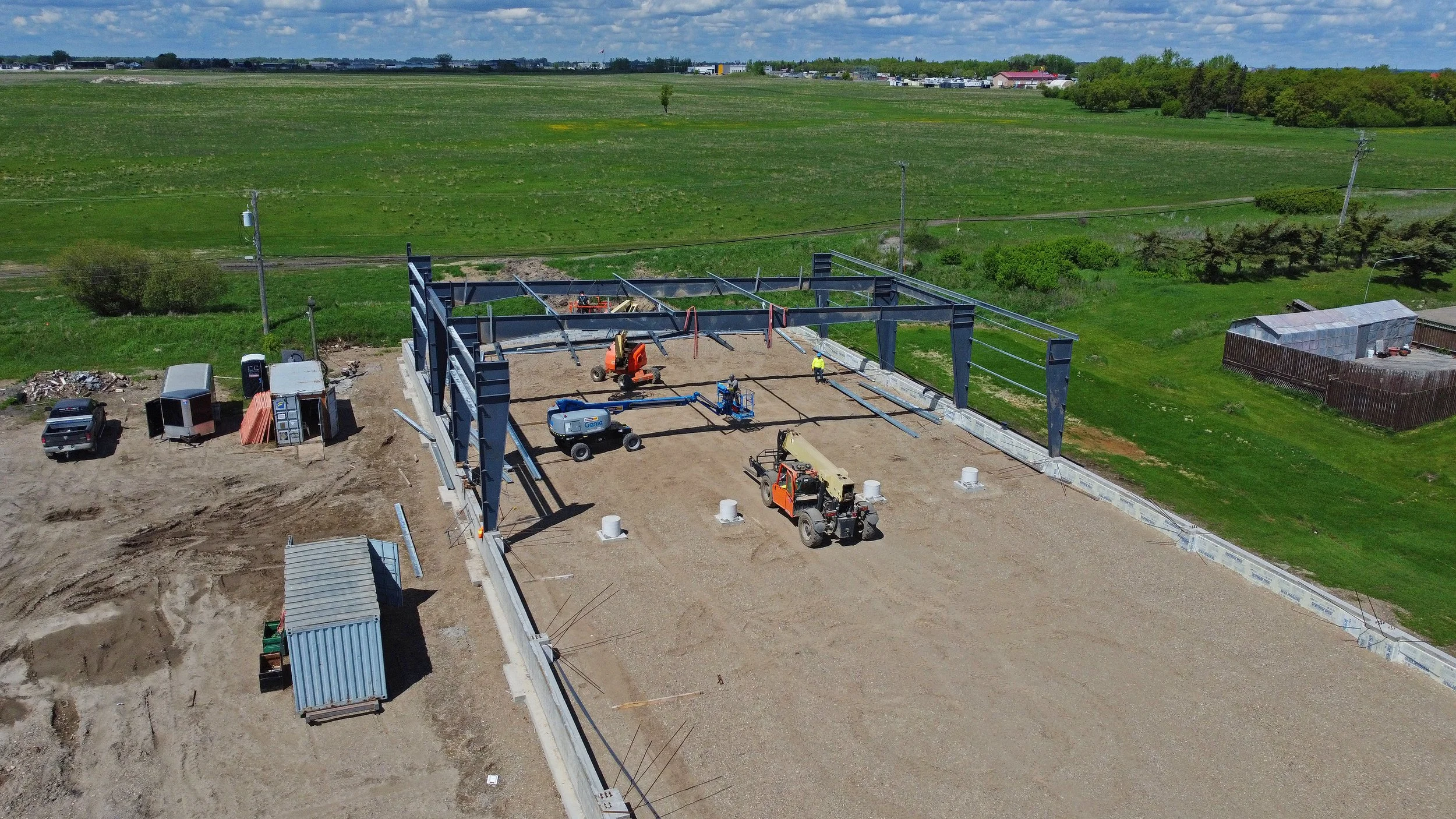 Construction site with steel framework, construction vehicles, and workers, set against green fields and blue skies.