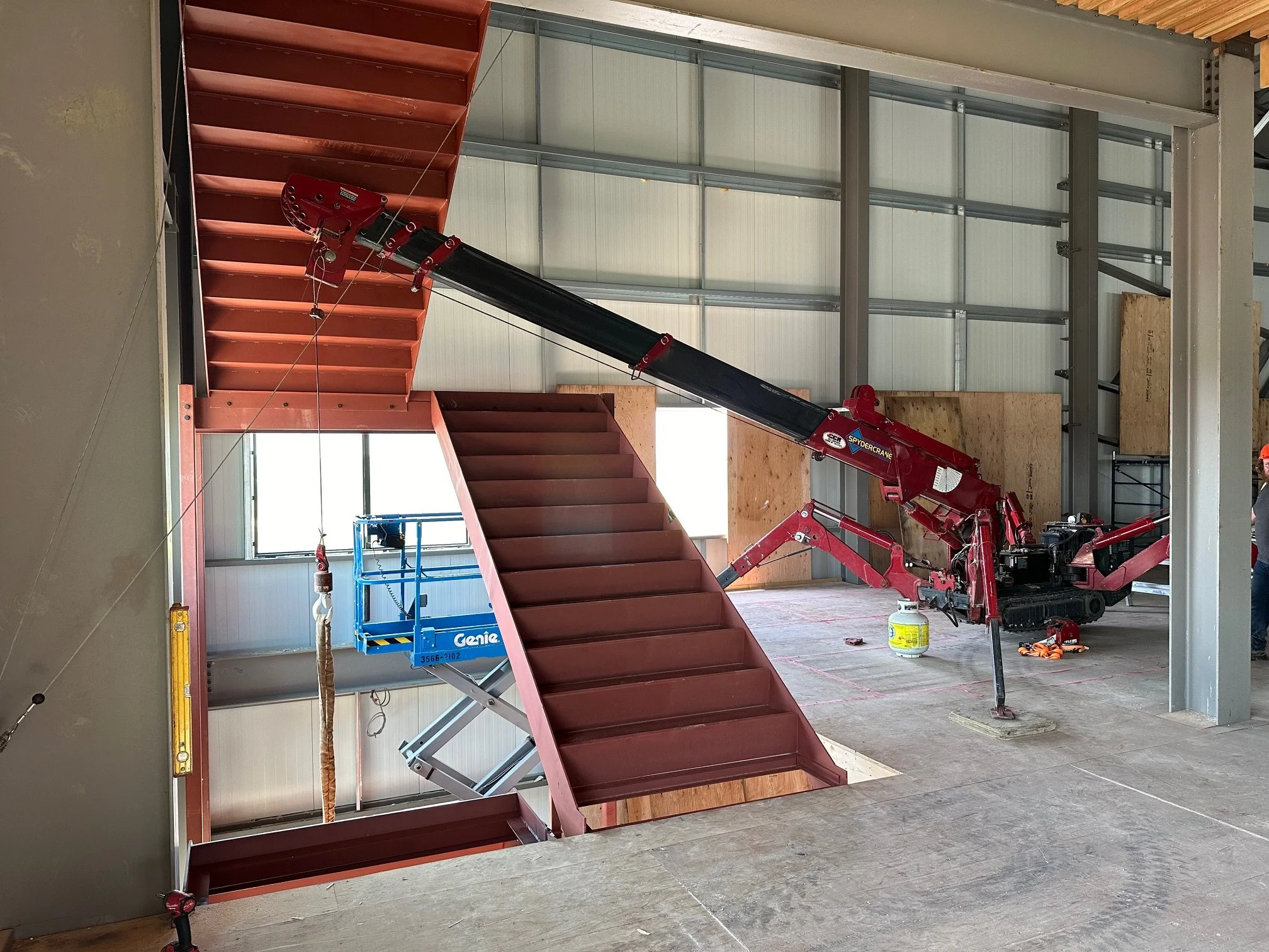 Interior of a building under construction with red metal staircase, a compact red crane, a blue lift platform, and unfinished walls.
