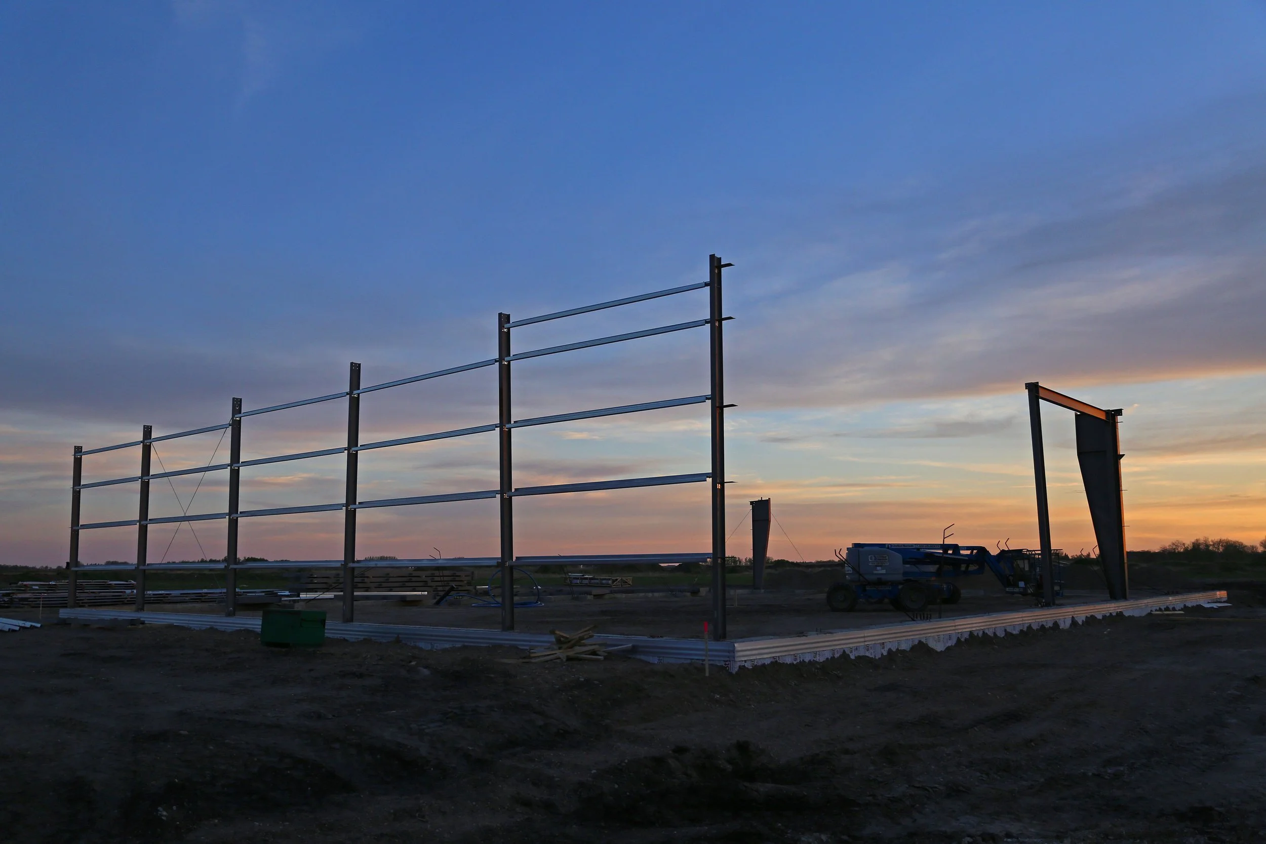 Steel framework of a building under construction at sunset, with construction equipment and a partly cloudy sky.