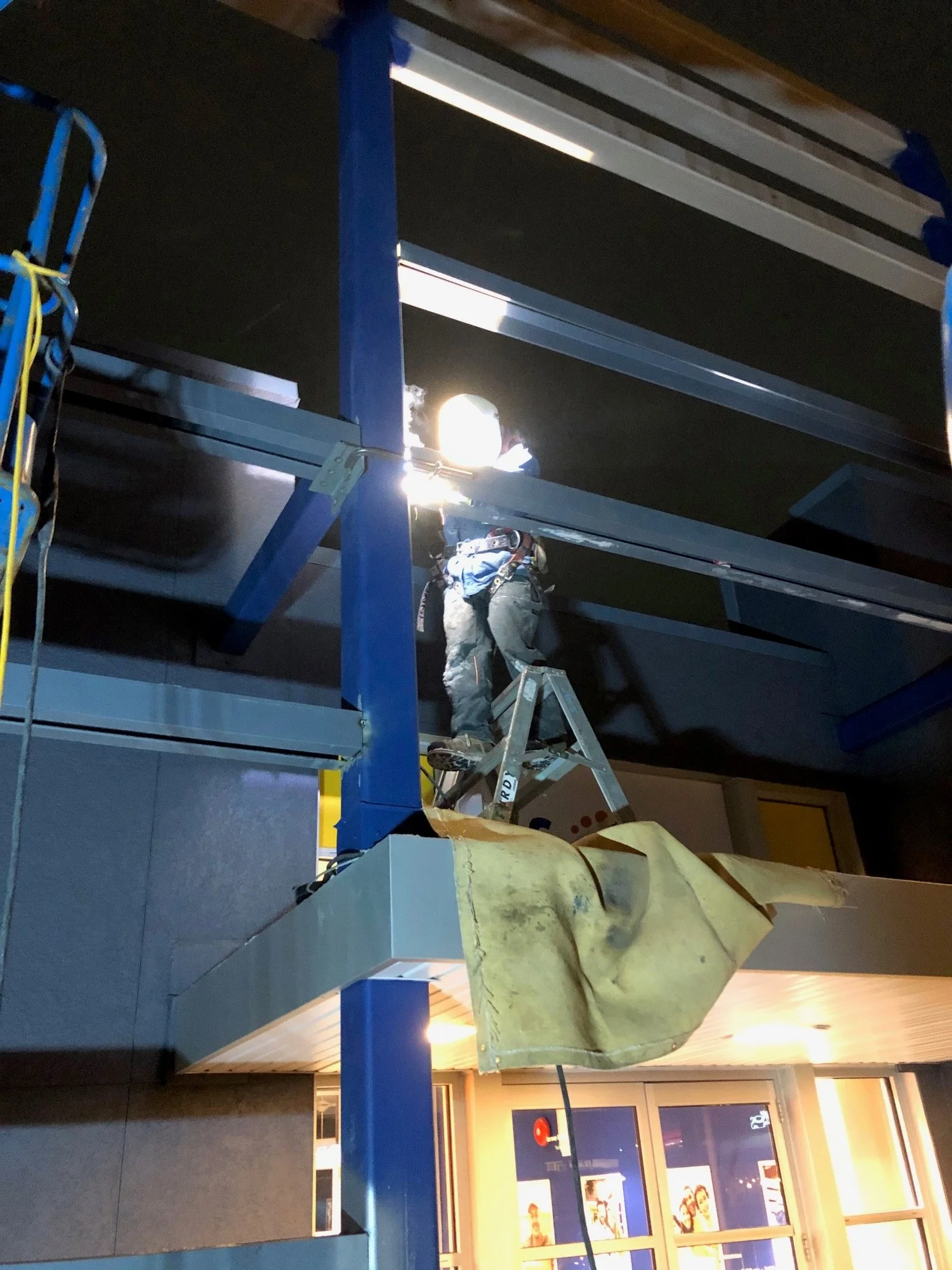 Worker standing on a ladder installing or repairing ceiling panels in an indoor commercial space, with tools and yellow protective covering nearby, illuminated by bright light.