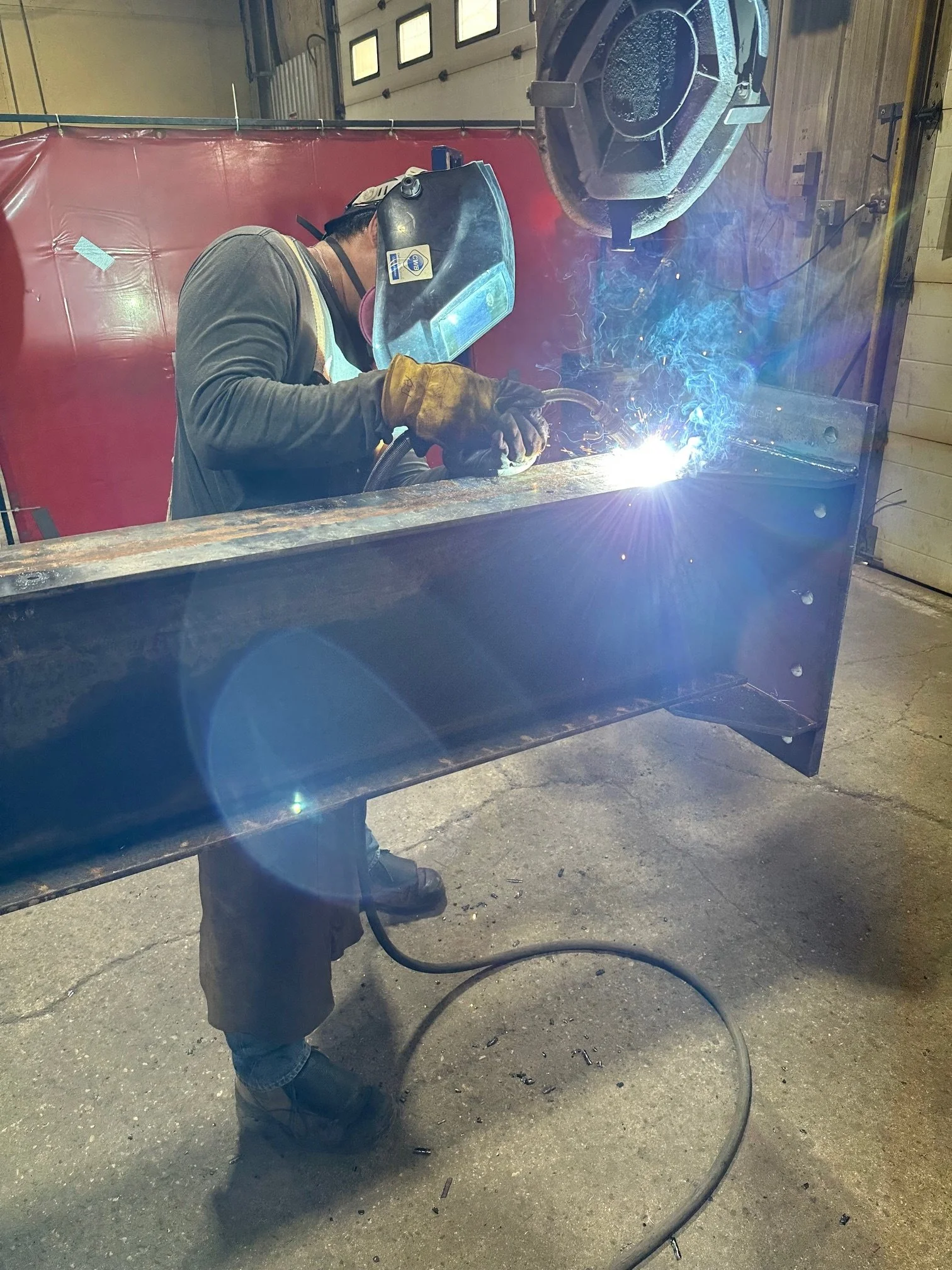 A person welding a large metal beam inside a workshop, wearing protective gear including a welding helmet and gloves, with sparks and bright light from the welding process.