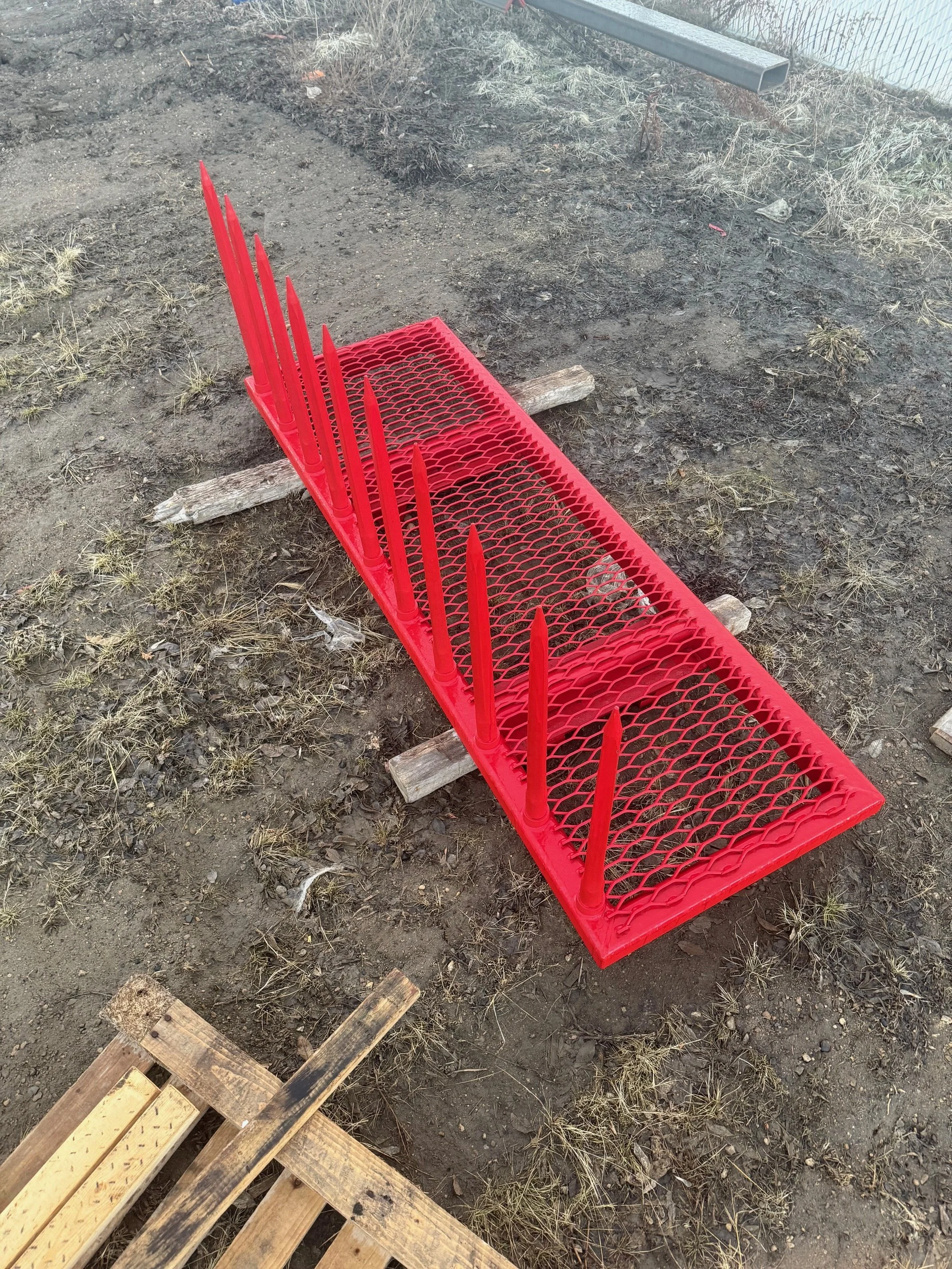 A red metal derby or obstacle course panel with spikes, laid on the ground in a dirt yard.