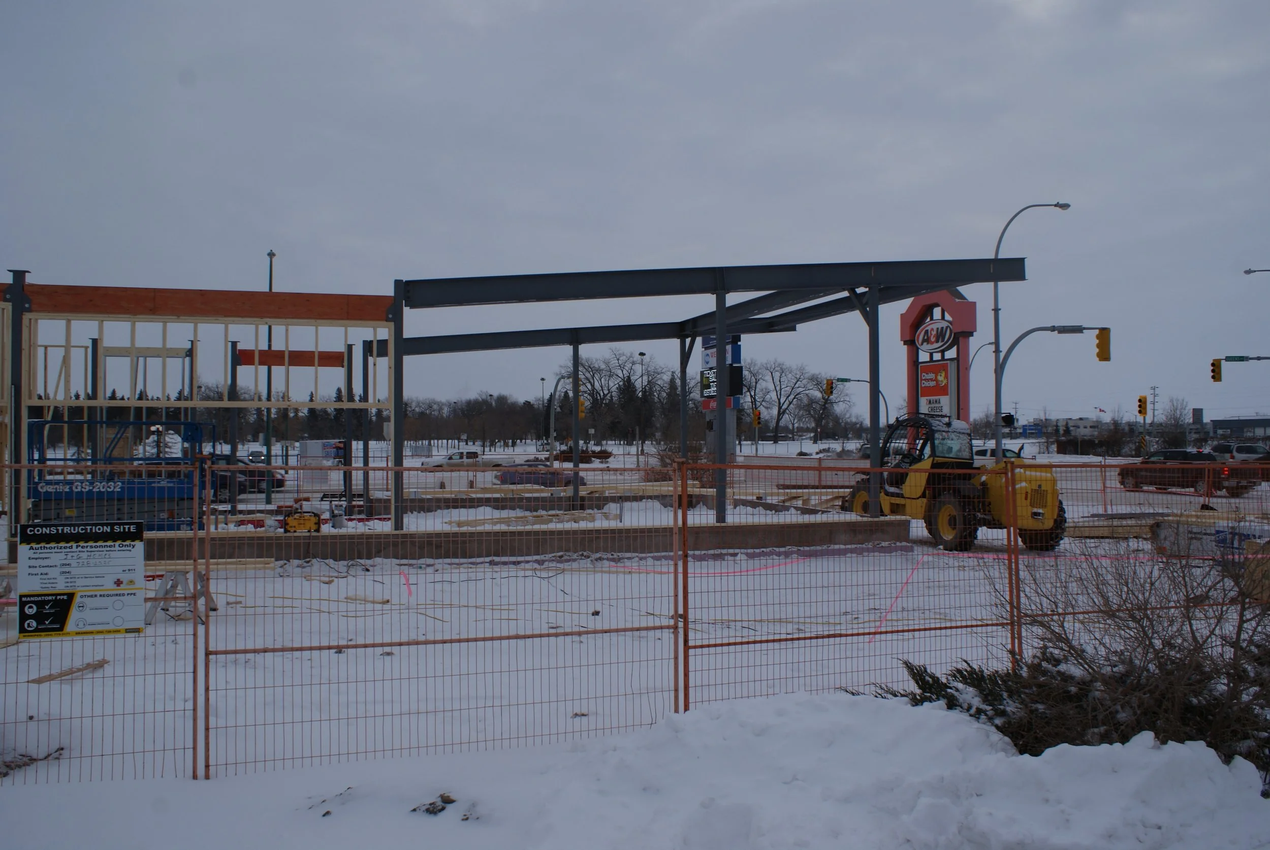 Construction site with a partially built structure, a small excavator, and snow on the ground, surrounded by orange fencing and streetlights in the background.