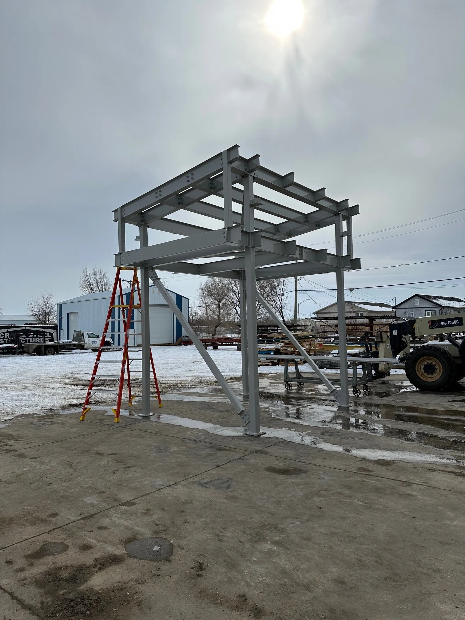 Metal structure under construction with a red ladder nearby, in an outdoor industrial area with snow on the ground and a cloudy sky.