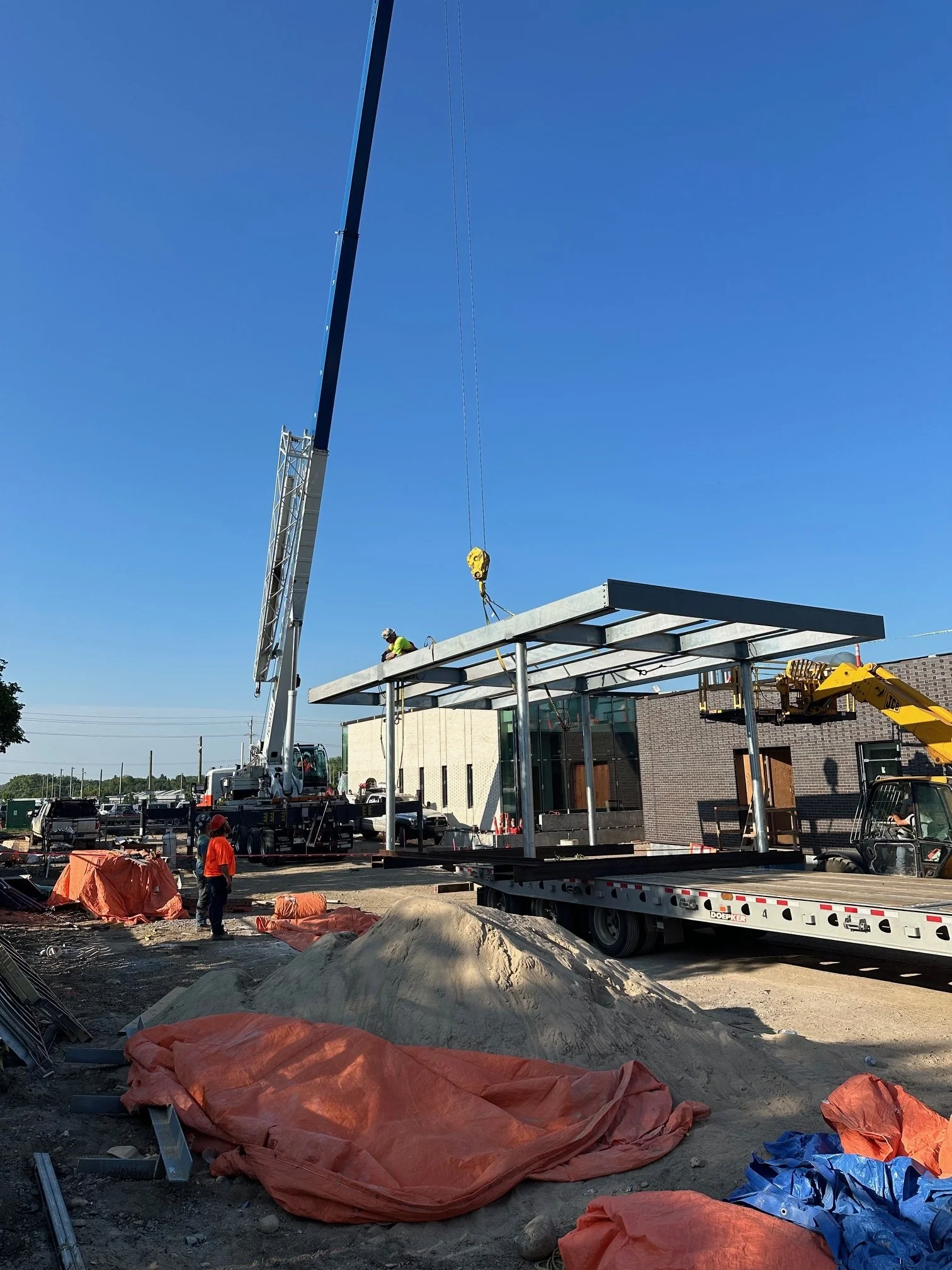 Construction workers and equipment at a building site with a crane lifting a large metal beam, blue sky in the background.