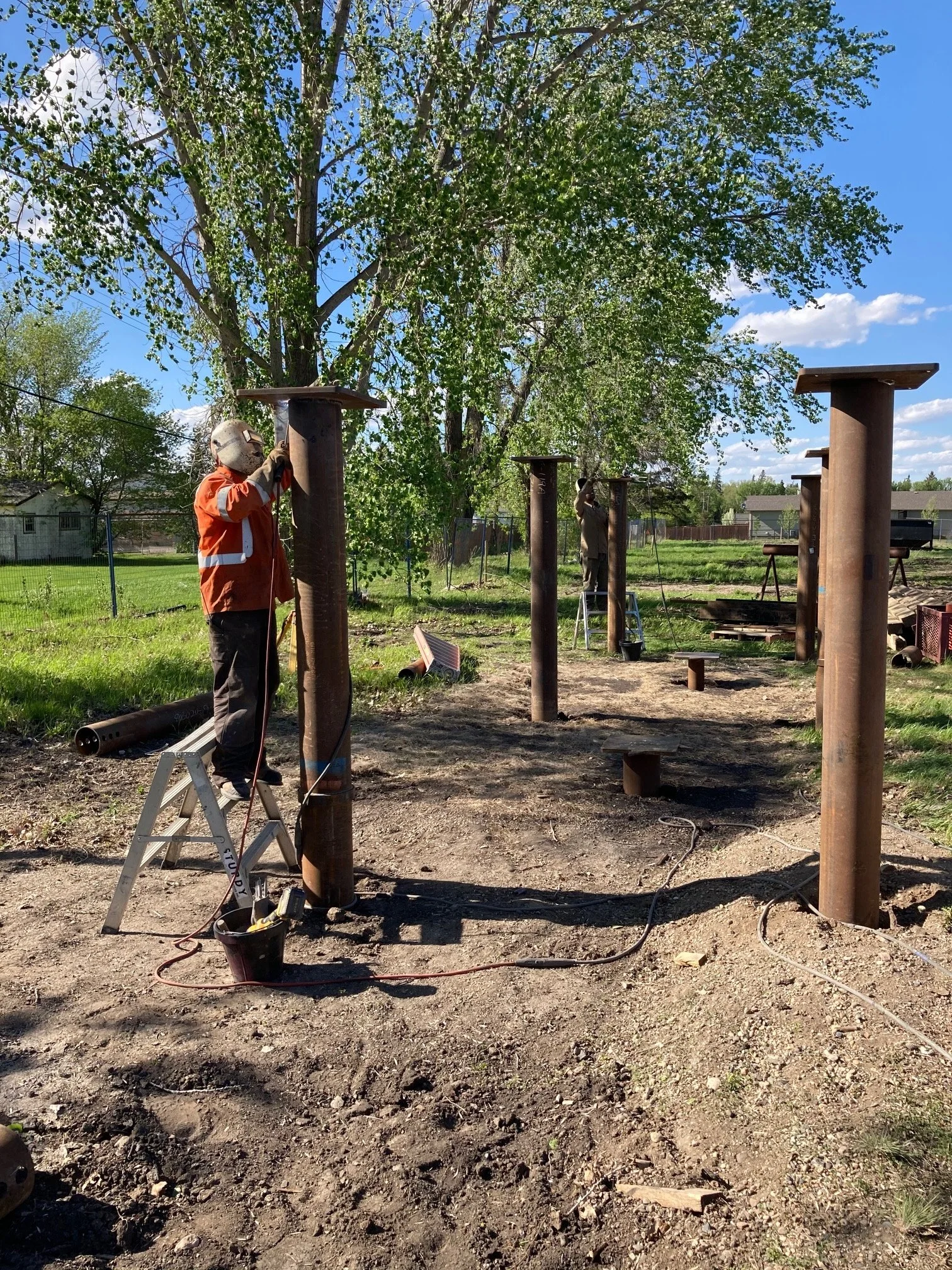 Two workers install metal poles with flat tops in an outdoor park area with grass, trees, and a dirt ground on a sunny day.