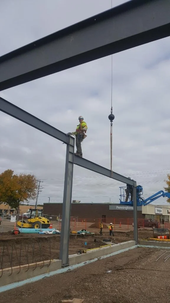 Construction worker in safety gear standing on a steel beam at a construction site, with a crane hook hanging nearby, and other workers and vehicles in the background.