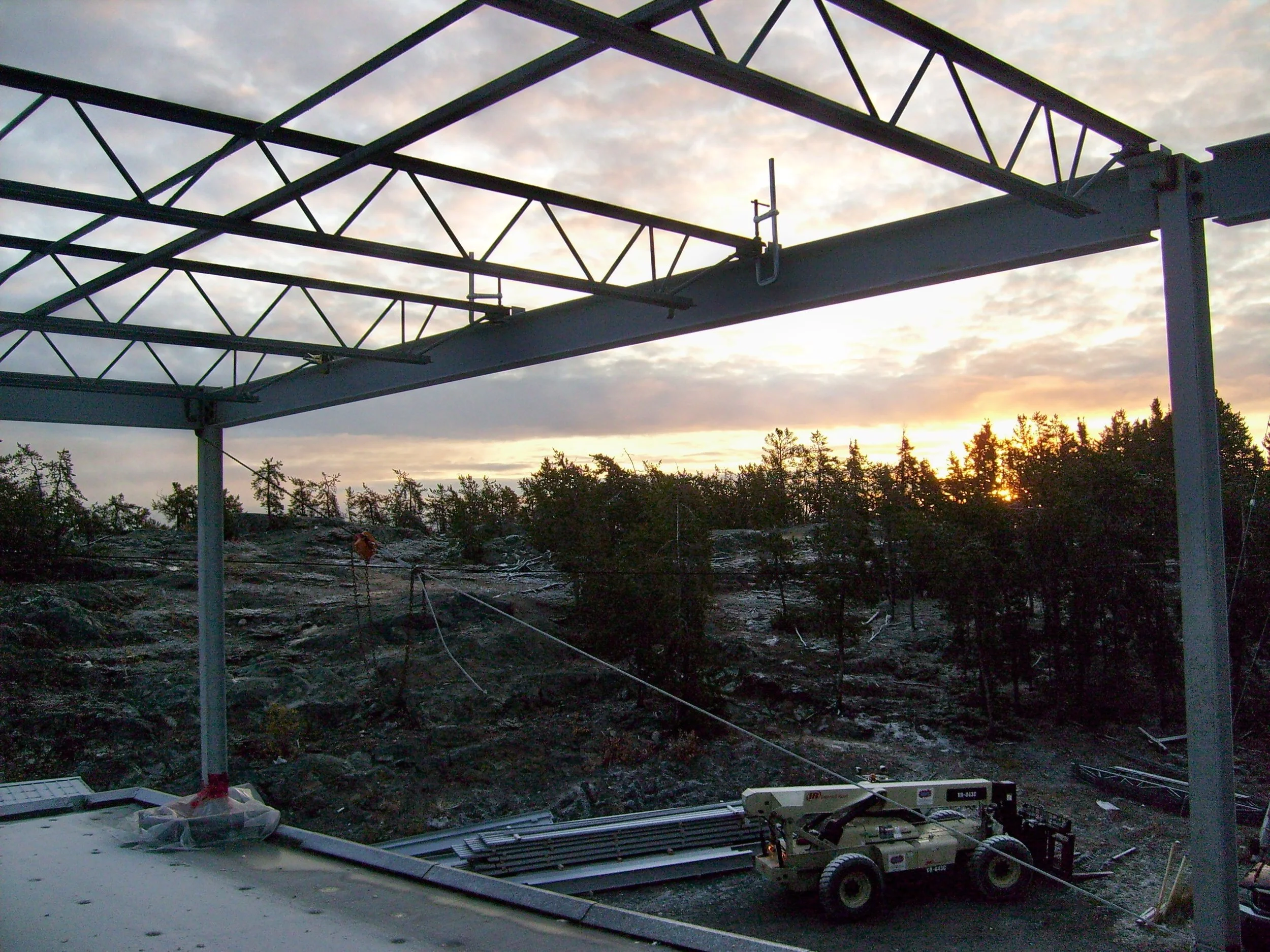 Construction site featuring a metal framework, a scissor lift, and a forested area at sunset.