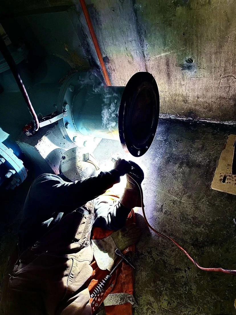 Worker welding metal pipe in a workshop at night, with sparks and smoke visible.