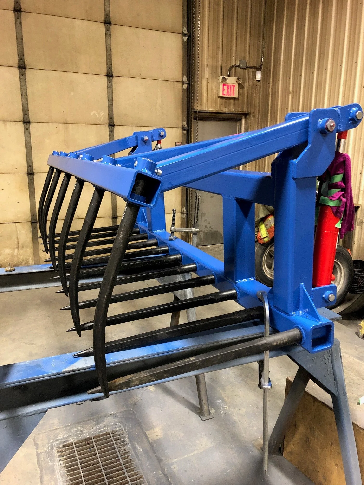 Blue hydraulic grapple attachment for heavy equipment, mounted on a metal stand inside a workshop.
