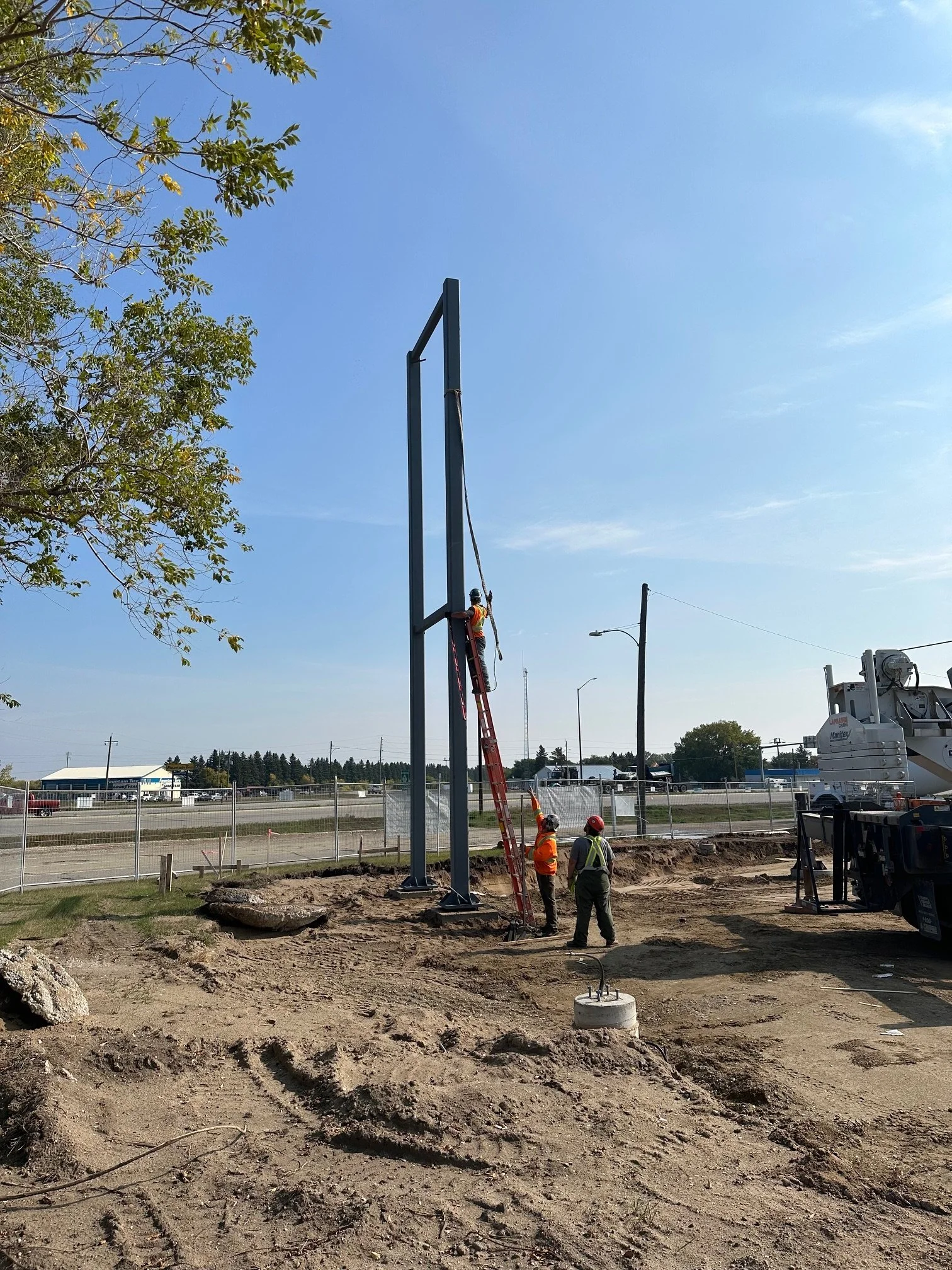 Construction workers installing a large billboard frame at a construction site on a sunny day, with a blue sky and sparse trees in the background.