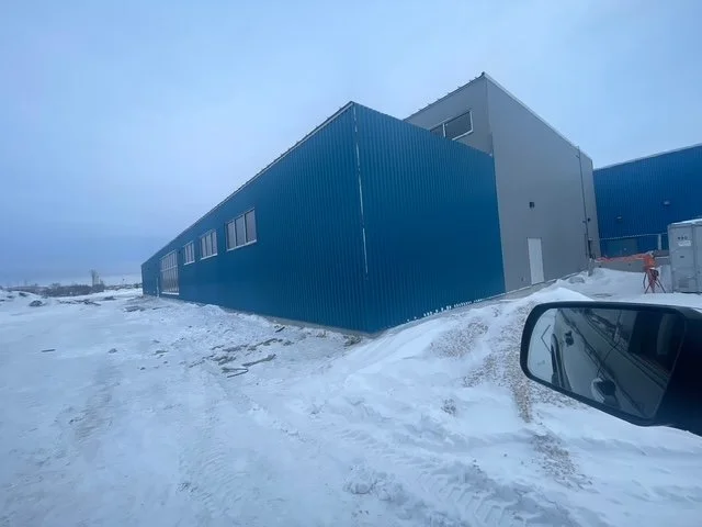 Large blue industrial building with a gray upper section, snowy ground, and a vehicle side mirror in the foreground.