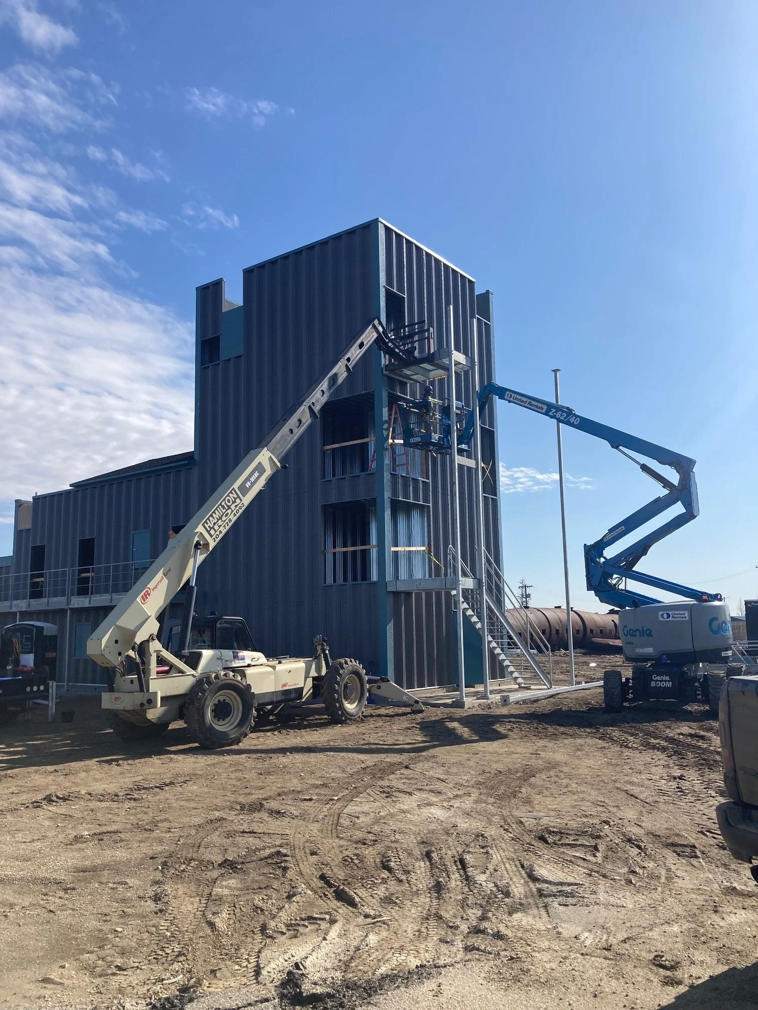 Workers on lifts installing or repairing windows on a modern multi-story building with a blue exterior, on a clear day with a dirt ground in the foreground.