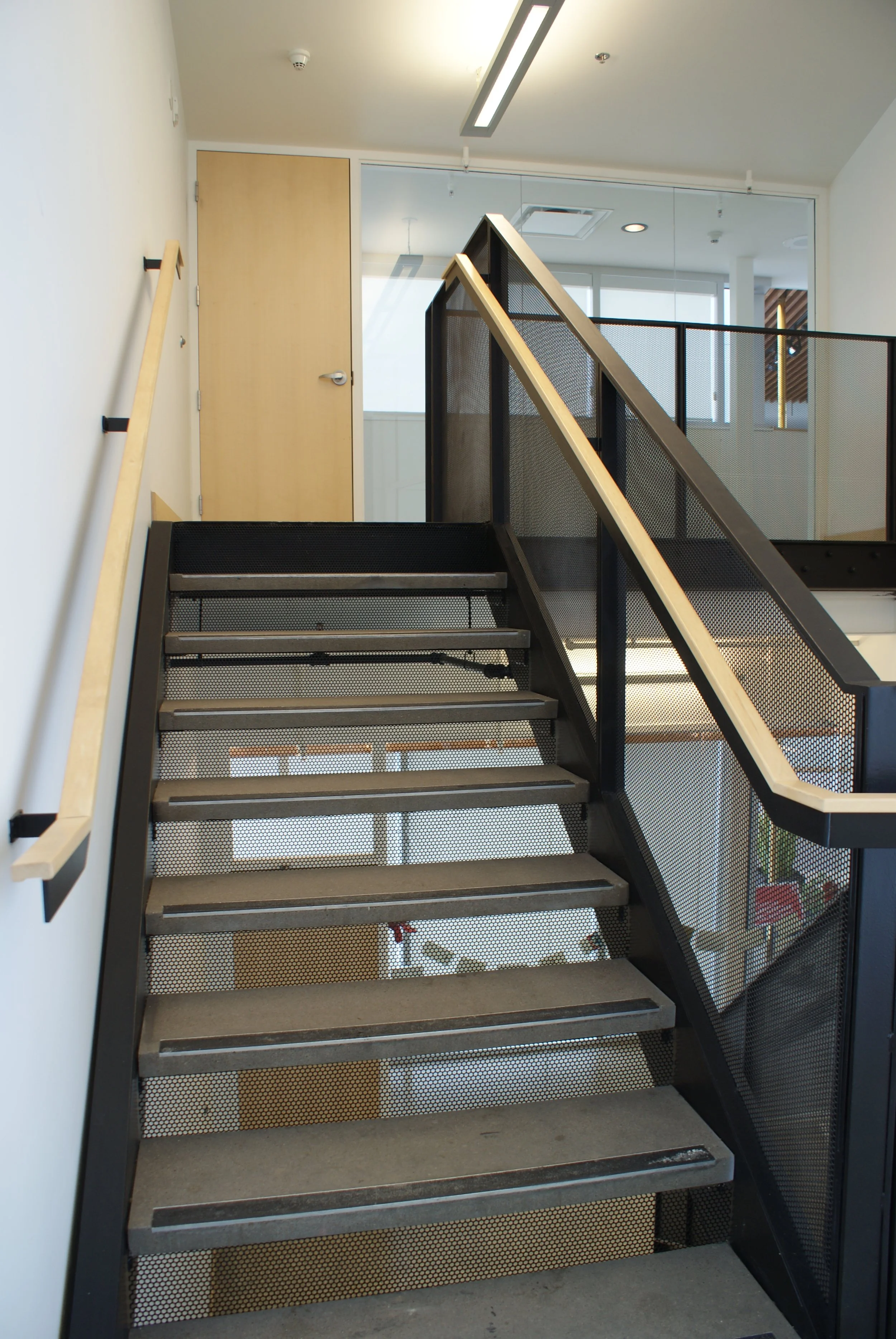 Interior view of a modern staircase with metal steps and mesh sides, leading up to a closed wooden door in a building.