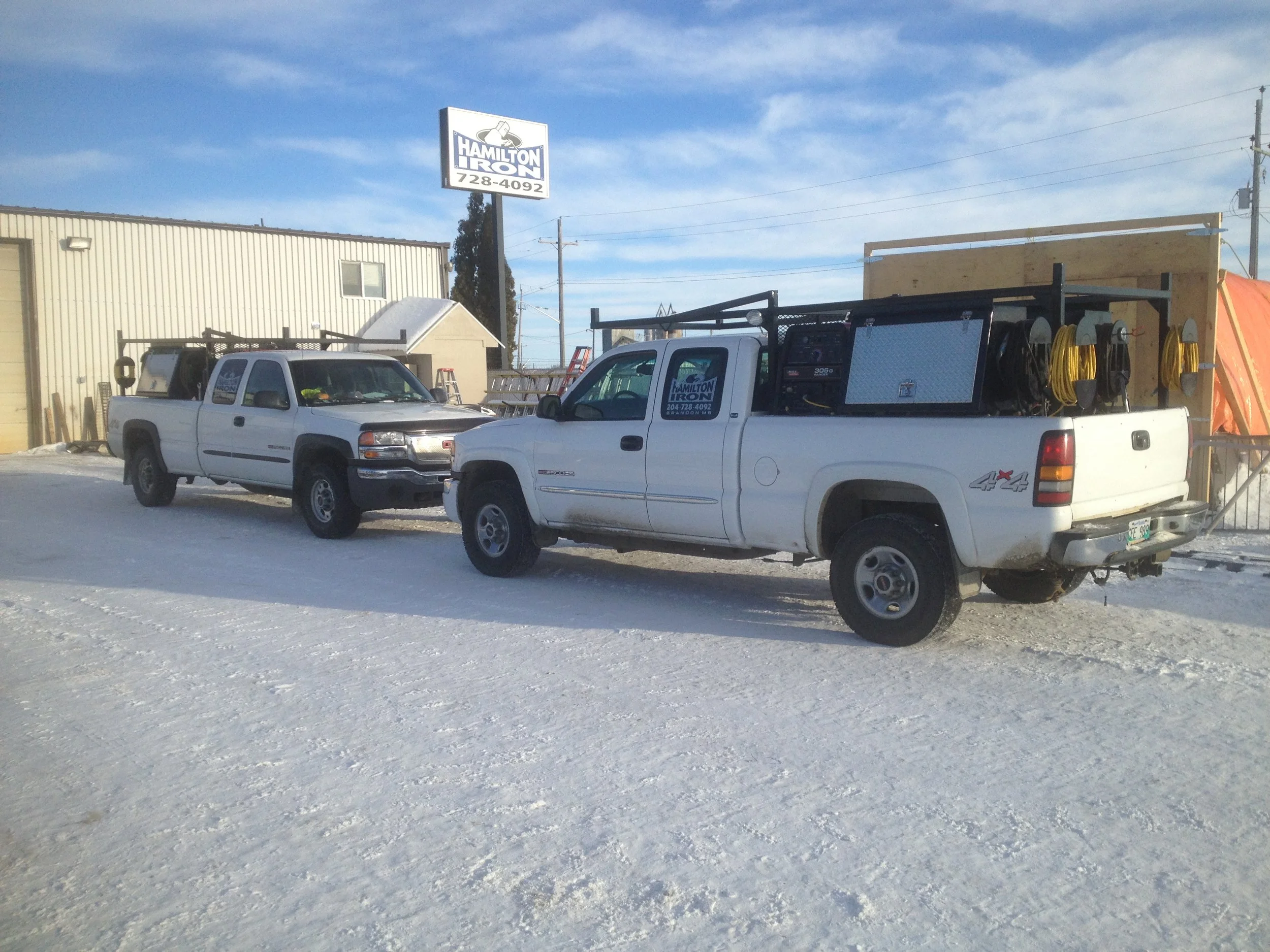 Two white pickup trucks parked on snowy ground outside a building, with a sign that reads 'Hamilton Iron' in the background.