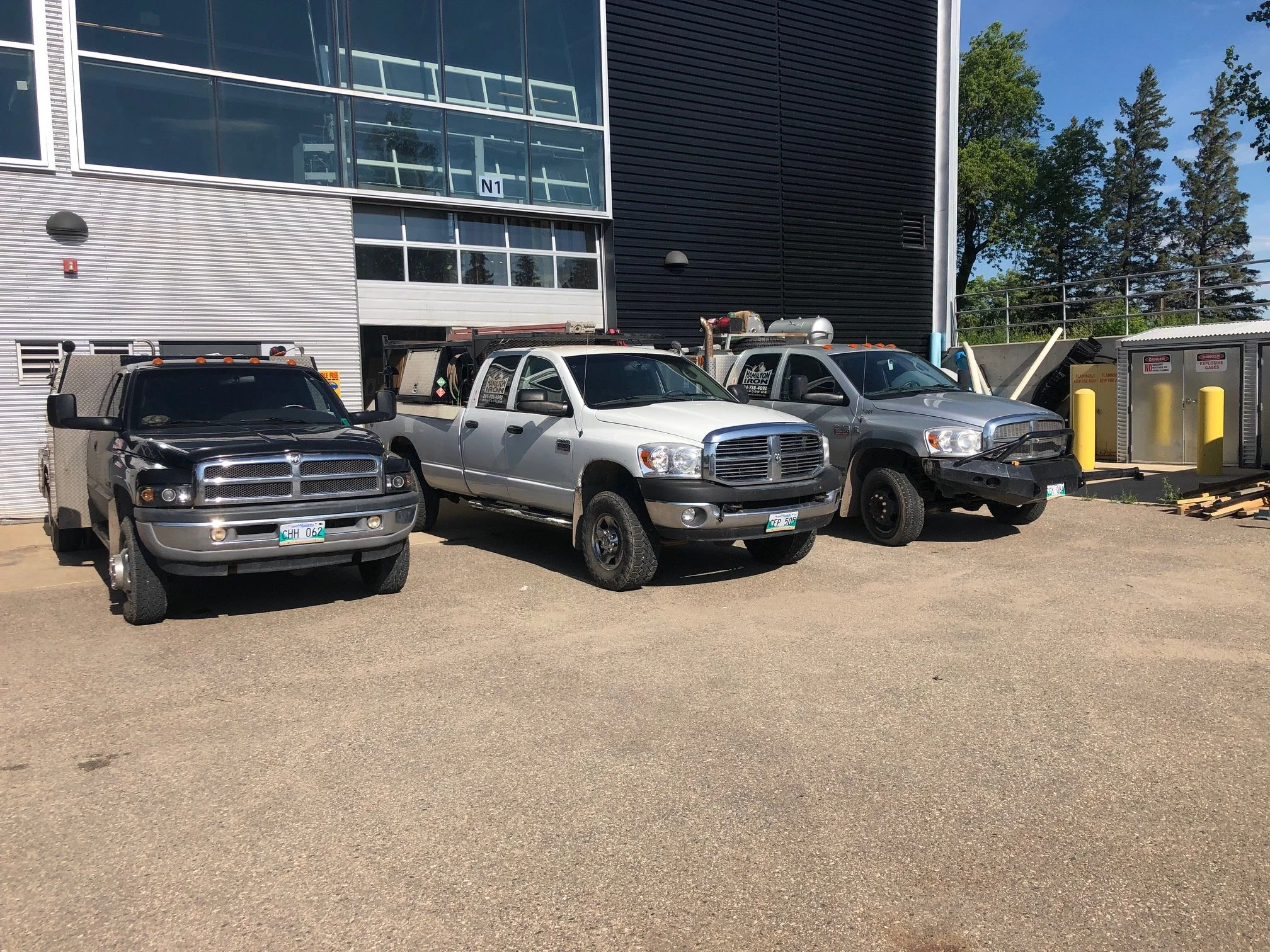 Three service trucks parked outside a modern building with gray and black siding, with trees and a clear blue sky in the background.