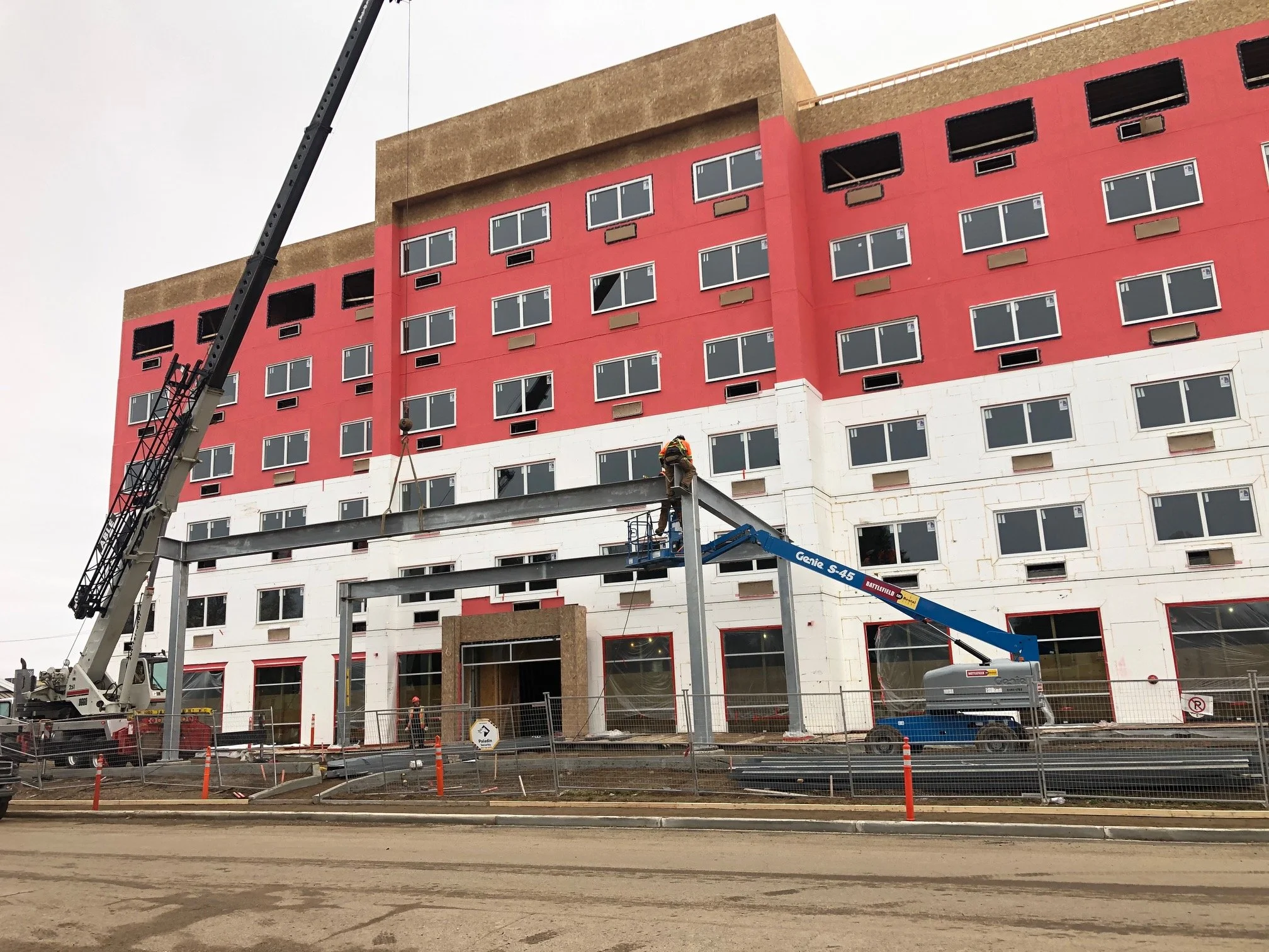 Construction workers on a lift installing structural beams on a multi-story building with a colorful facade, surrounded by construction fencing and equipment.