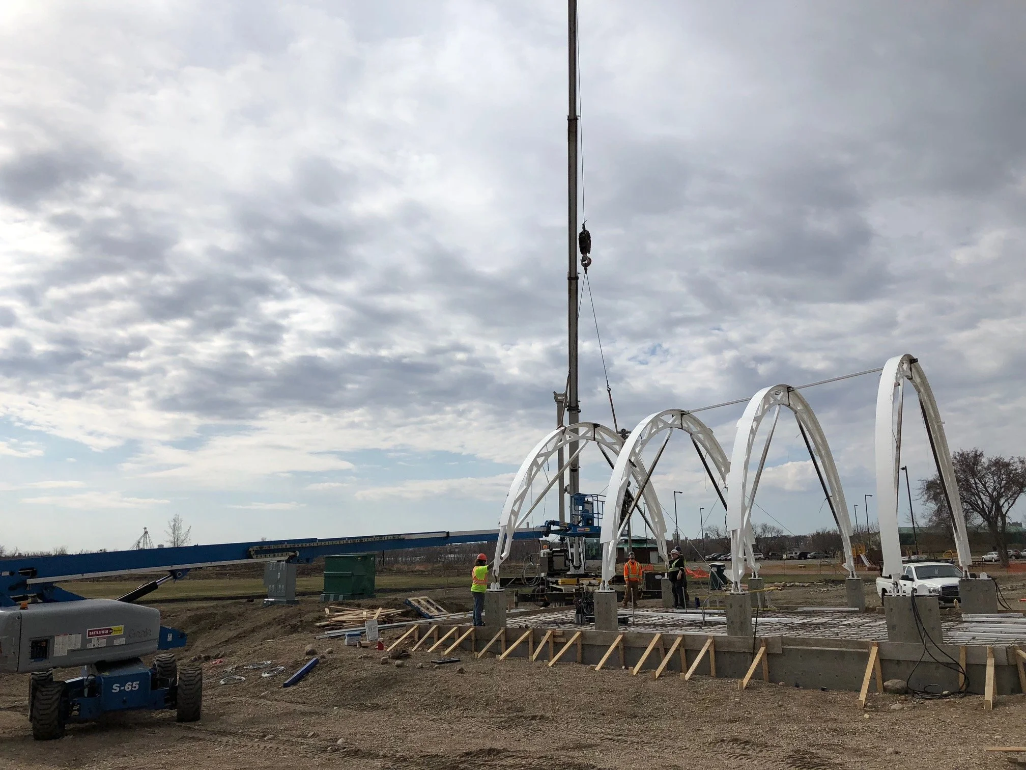 Construction workers and equipment installing a large, white arch structure at a construction site on a cloudy day.