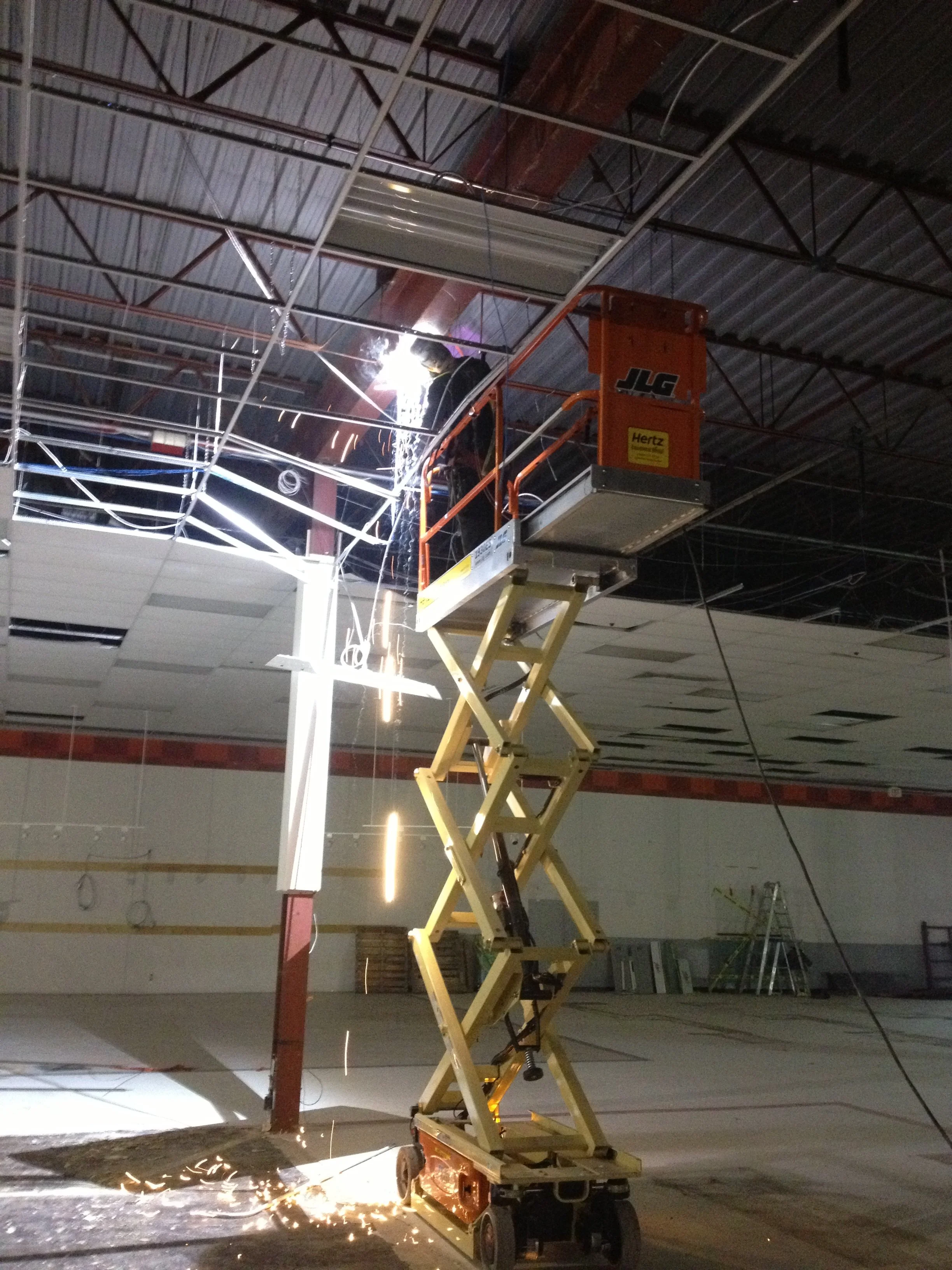 A worker welding in an orange scissor lift inside a building, sparks flying as metal is being welded to a metal framework ceiling.