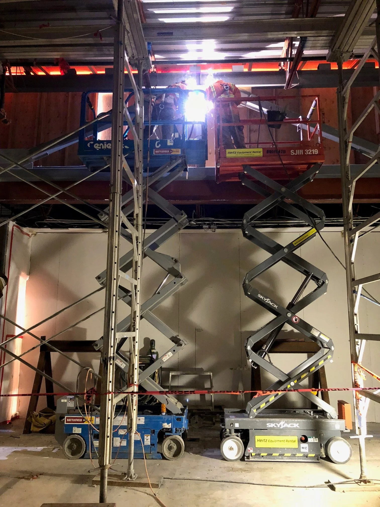 Construction workers in safety gear working on an upper level using scissor lifts in a building under construction.