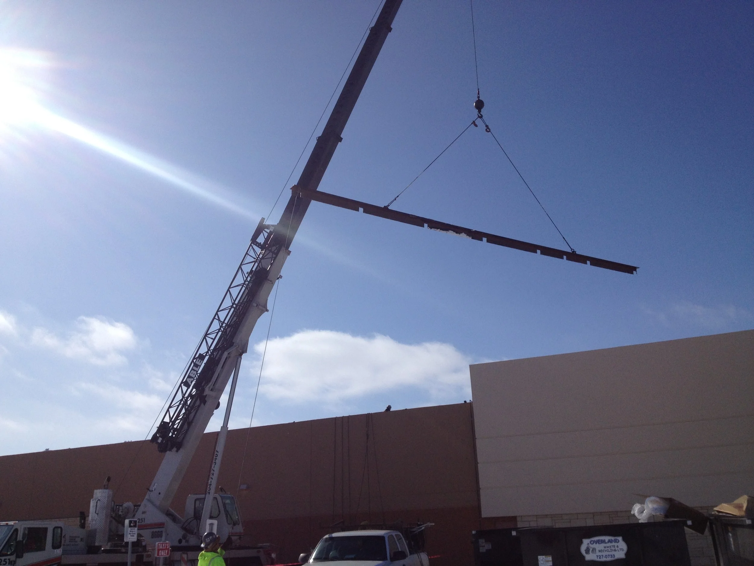 A crane lifting a long steel beam near a building at a construction site on a sunny day.