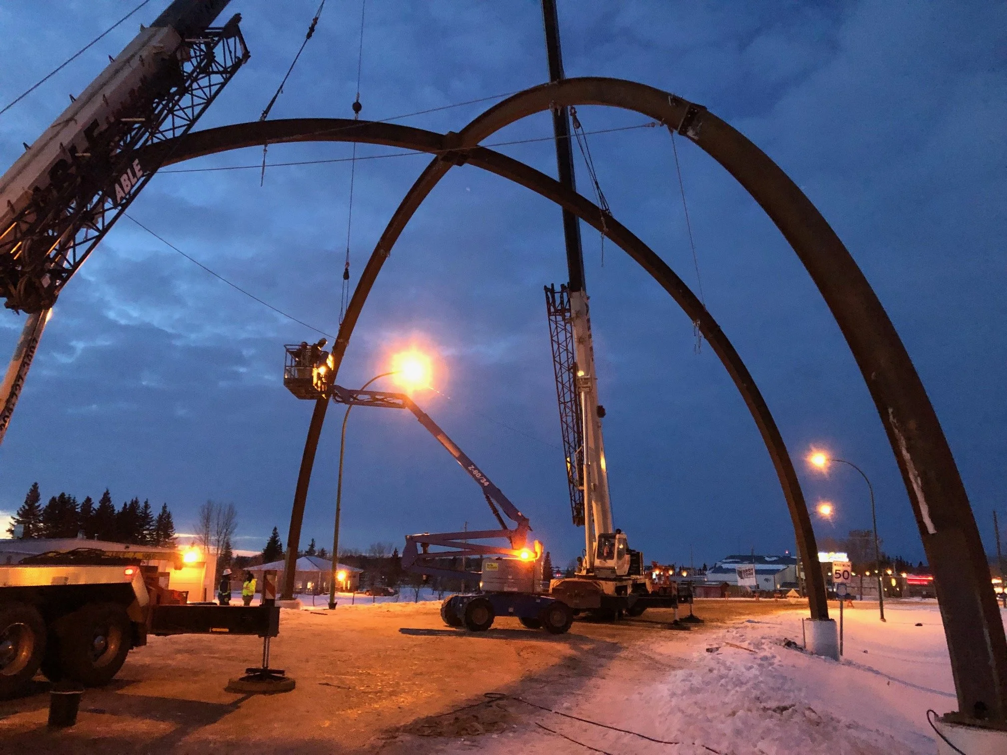 Nighttime construction scene with large metal arches being assembled, construction workers, and cranes working under streetlights with snow-covered ground.