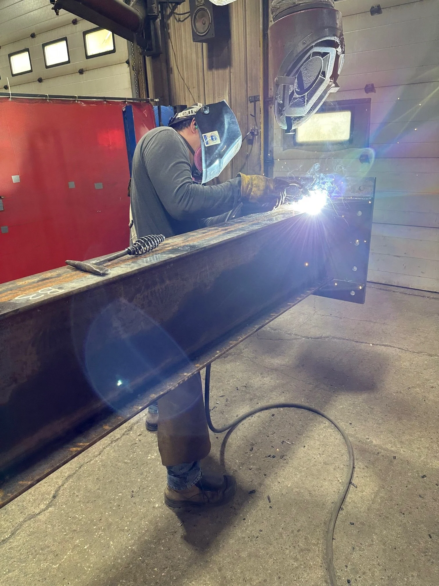 A person welding a large metal beam in a workshop, wearing protective gear including a welding helmet and gloves, with bright sparks and light from the welding process.