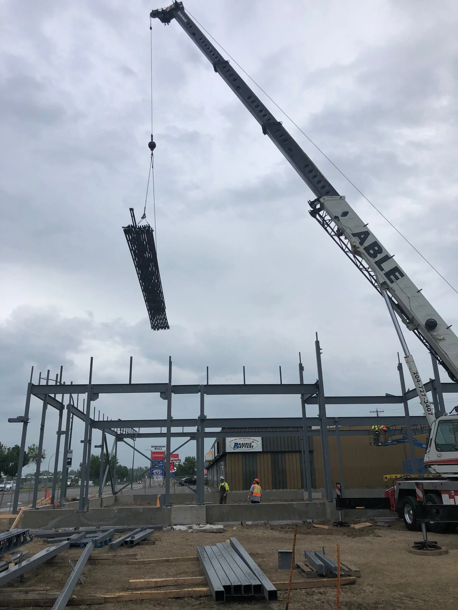 Construction site with a crane lifting a steel frame, workers in safety vests and helmets, and a partially built steel structure.
