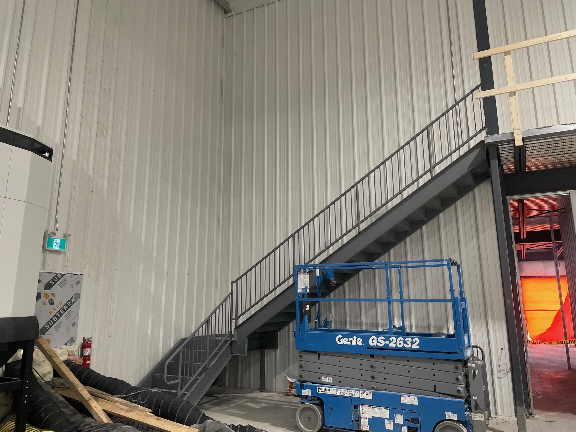 Industrial warehouse with metal stairs and a blue Genie scissor lift, construction materials and equipment nearby, and a red safety curtain at the entrance.