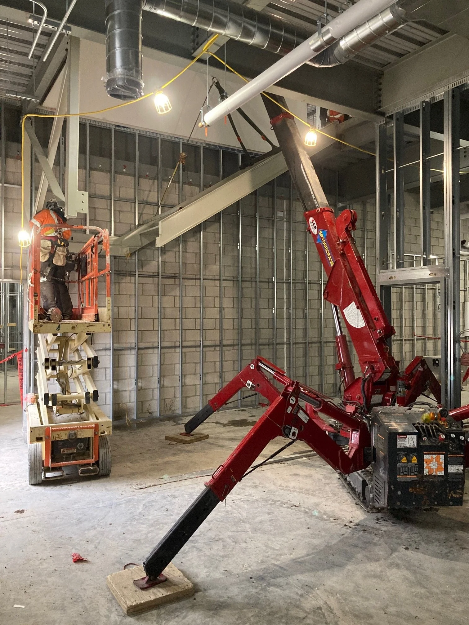 Construction site with a worker on a lift and a large red crane installing structural beams in a building's framework.