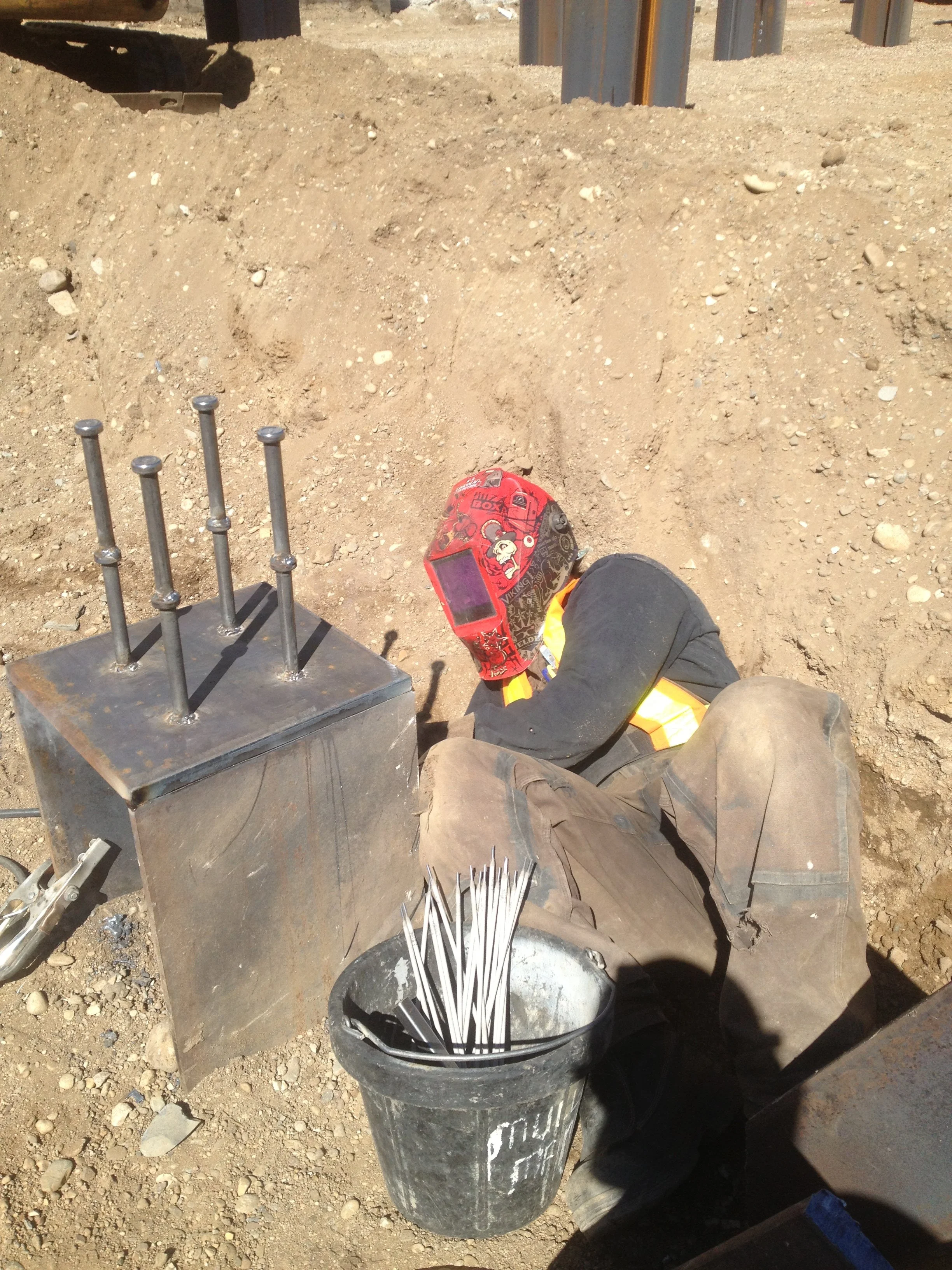 A construction worker wearing safety gear, including a helmet with a face shield, is sitting on the ground at a construction site. In front of them is a metal block with six nails standing upright and a black bucket filled with bent metal rods or reb