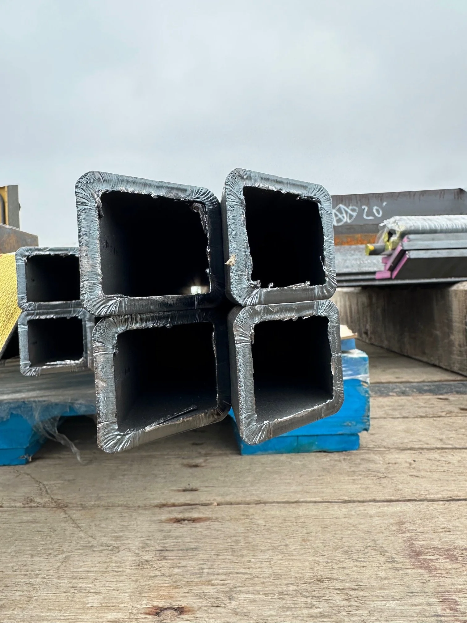 Stacked steel square tubes on a wooden surface with construction materials in the background.