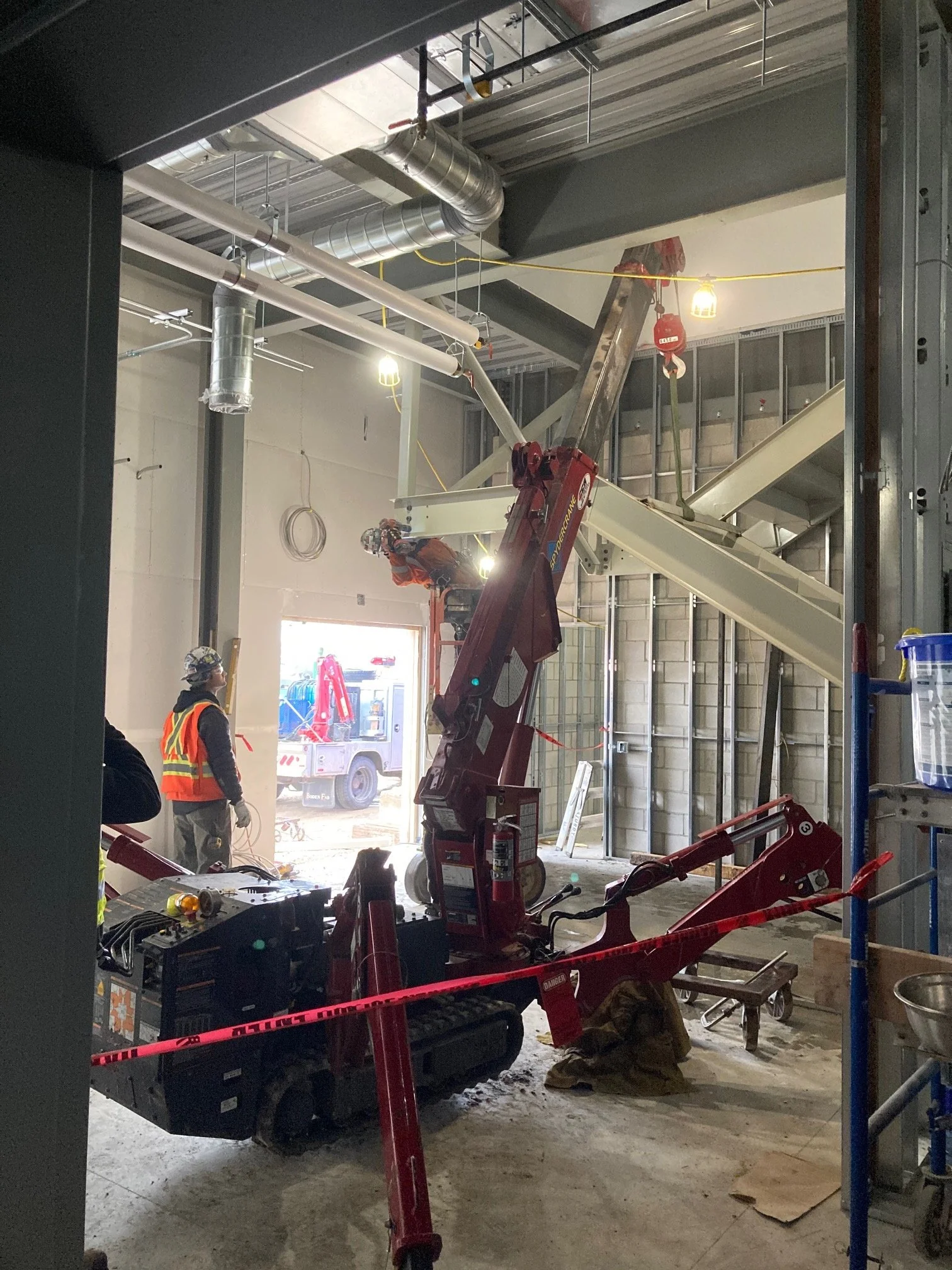 Construction workers inside a building under construction using a small crane to lift materials, with exposed ductwork, electrical wiring, and unfinished walls.